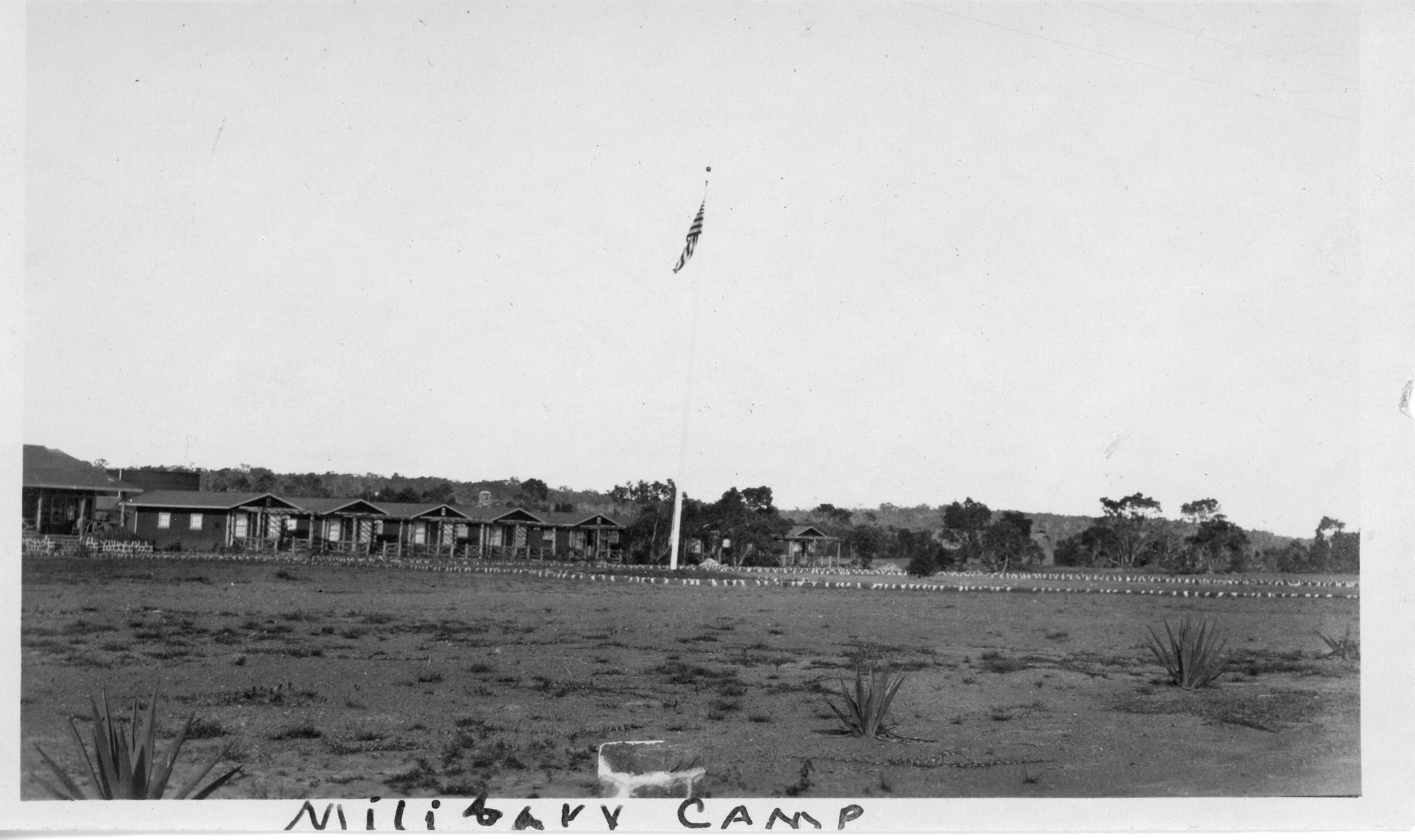 A black and white photo of the Kilauea Military Camp. This picture is a wide view of the camp with a flagpole in the middle of the image. The flag on the pole is moving with the wind. The picture shows multiple buildings in the distance lined up next to each other for housing the military coming to Hawaii. Behind the buildings are large trees. The ground seems to be dirt with a few grass spots and cacti scattered around. The photo has the caption "Military Camp" written on the bottom.