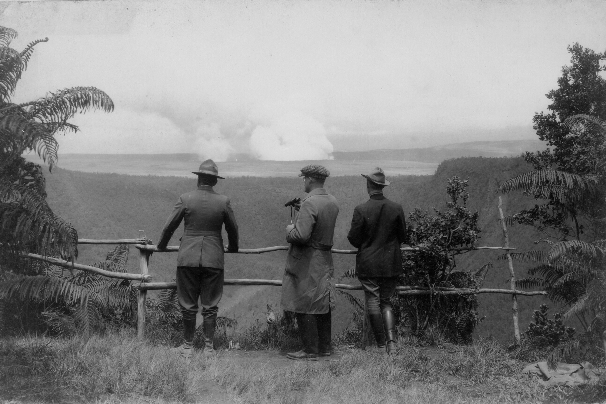 Black and white photograph of three unidentified men standing at a rustic wooden railing that overlooks a vast valley. The men are National Park Service uniforms. The man in the middle holds a pair of binoculars in his right hand. Lush vegetation is present all around them. Beyond the railing, there is a expansive valley below. The valley sits among tall elevation with dense vegetation. In the distance, smooth, flat land can be seen in which two large smoke clouds are emerging from the ground. The smoke appears to be rising from the caldera.