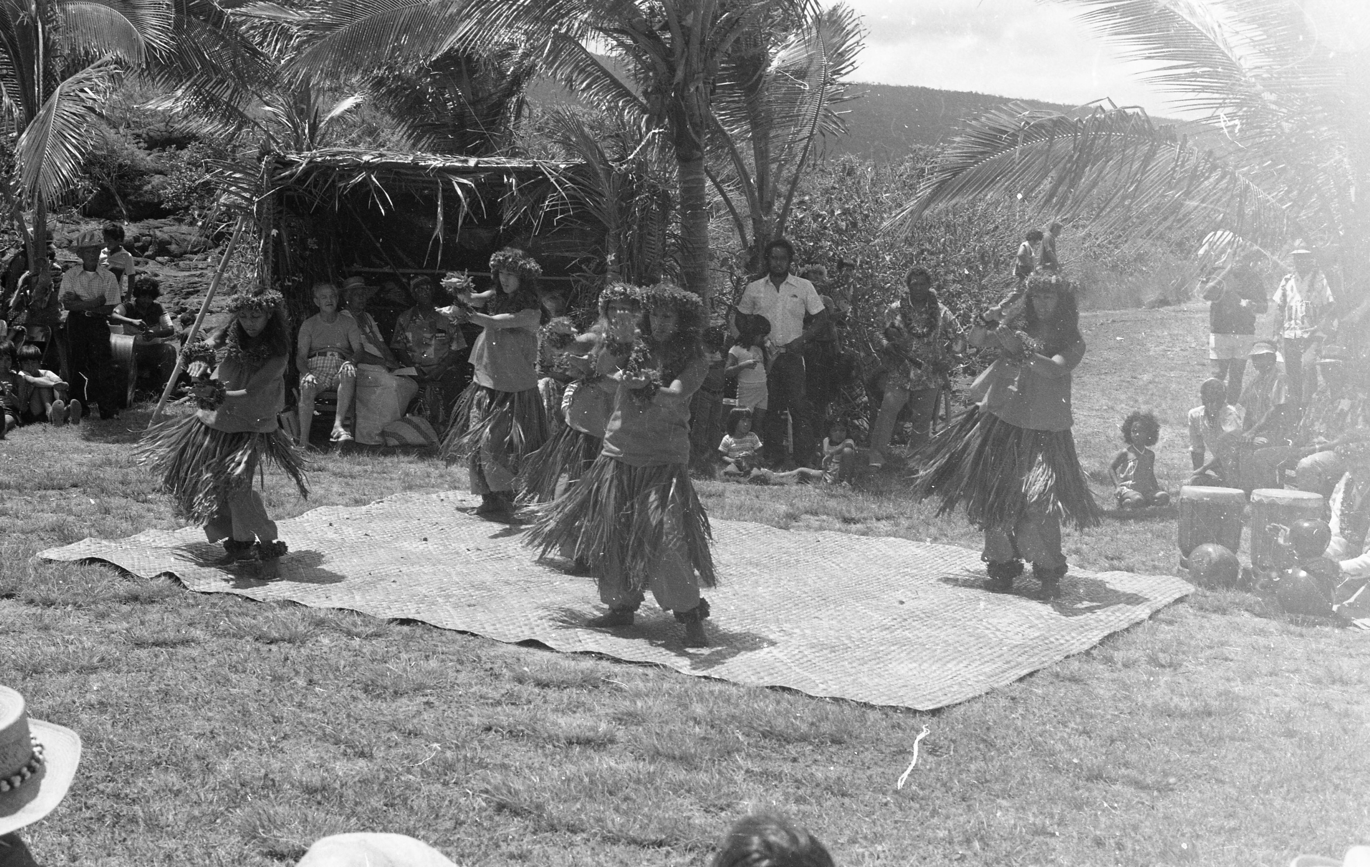 A black and white image of five hula dancers performing on a woven mat. The women are located in the center of the image standing on a large woven mat. Each of the women are wearing a floral headband, leis, floral bracelets, one on each wrist, a tube top, a grass skirt with pants underneath, and a floral anklet on each of their feet. Their arms are stretched out in front of them with their right arm hoovering over the left. In the background there are people sitting on the ground, in chairs, and standing watching the performers. Some of the people are seen sitting inside of a booth towards the top left side of the image.