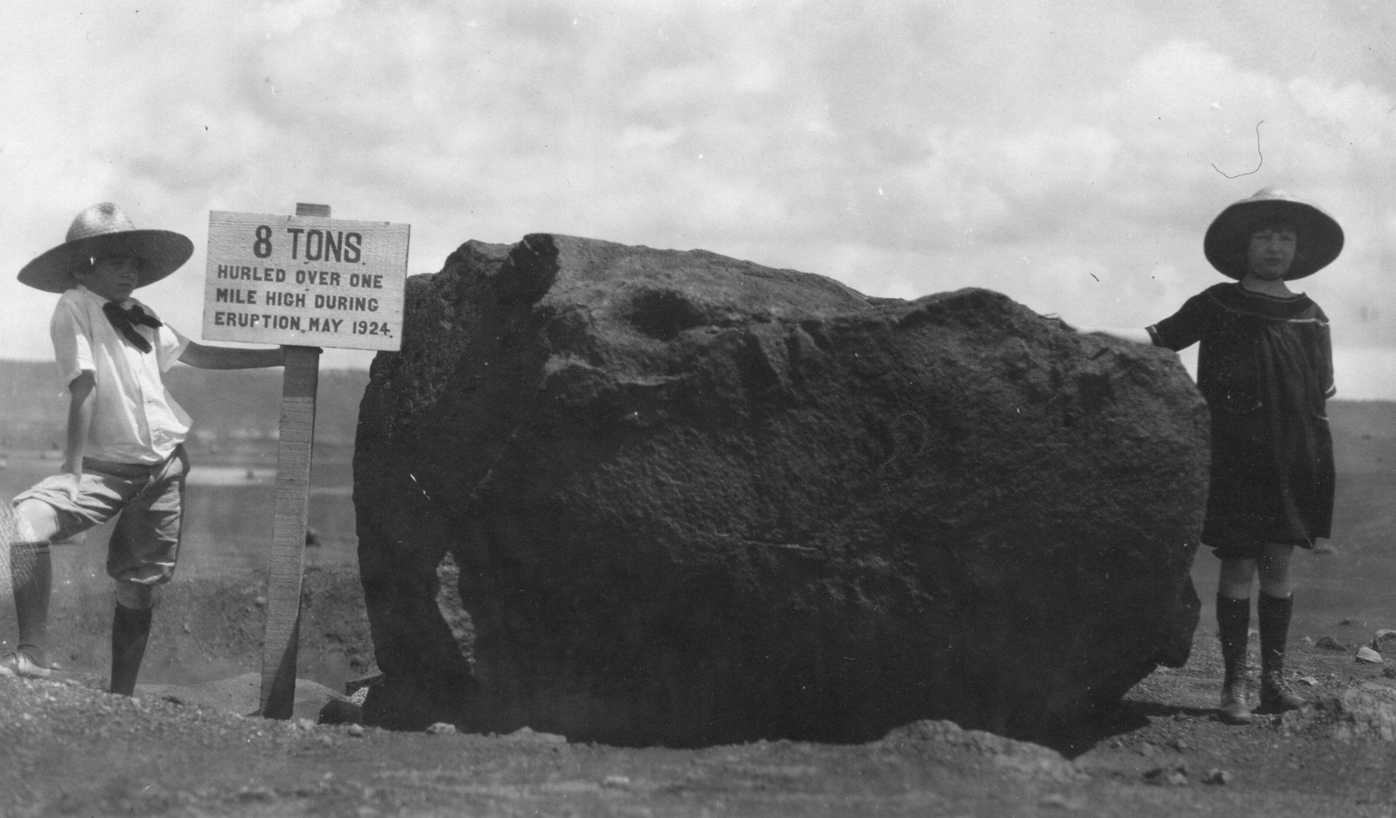 The image is in black and white. Two girls, Charlotte Lovejoy and Margaret Boles, pose on either side of a large boulder hurled from Halema‘uma‘u  during an eruption. The rock fills the center of the image. On the left, Charlotte poses with an explanatory sign, her left arm extended to hold the back of the signpost while the other rests on her bent right knee, which is higher than her other leg. She is wearing a hat, button-up shirt, shorts, socks, and shoes. The sign she is holding onto, a light-colored sign with dark, all-capitals font, reads, "8 Tons. Hurled over one mile high during eruption, May 1924." Margaret, on the right side of the stone, is wearing a hat, dress, socks, and shoes. She has one arm resting on the boulder while the other is behind her back. The background is barren, ashy, and rocky. The sky is cloudy.