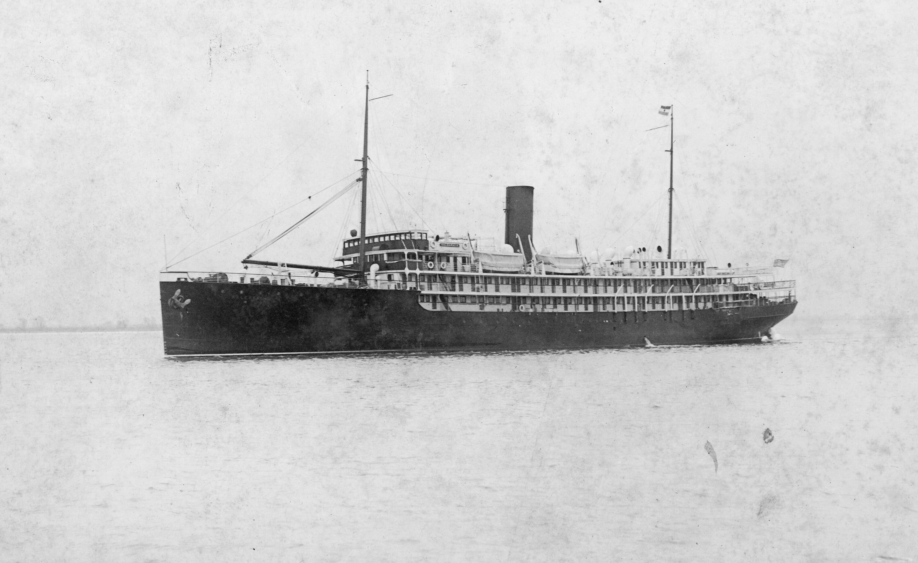 Black and white photograph of an unidentified passenger ship at sea. Ship is in the center of the photograph, sky and water are almost indistinguishable. A couple of people are visible near balconies. An American flag is hoisted at the stern.