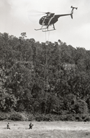 A black and white image of a helicopter in the sky with a rescue rope dropped. Towards the bottom left corner are two people walking towards the rope that is hanging from the helicopter in the center of the image.