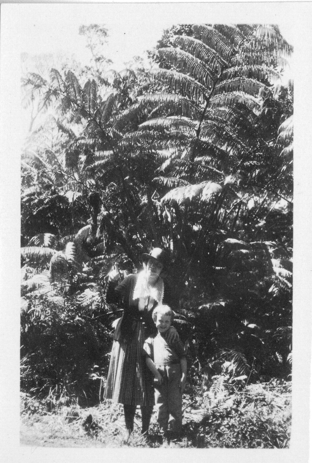 A black and white image of a woman and a boy standing in front of a hapuʻu or fern tree. The woman and boy are located in the center of the image. The woman is standing to the right of the boy. The woman is wearing a hat, blazer, a shirt with an exaggerated collar, and a crosshatch skirt. She has her left hand resting on top of the boy's left shoulder. Her right hand is pointing up, and she is looking towards the left side of the image. The boy is wearing a t-shirt and pants. He is smiling at the camera. Behind them is a large hapuʻu.