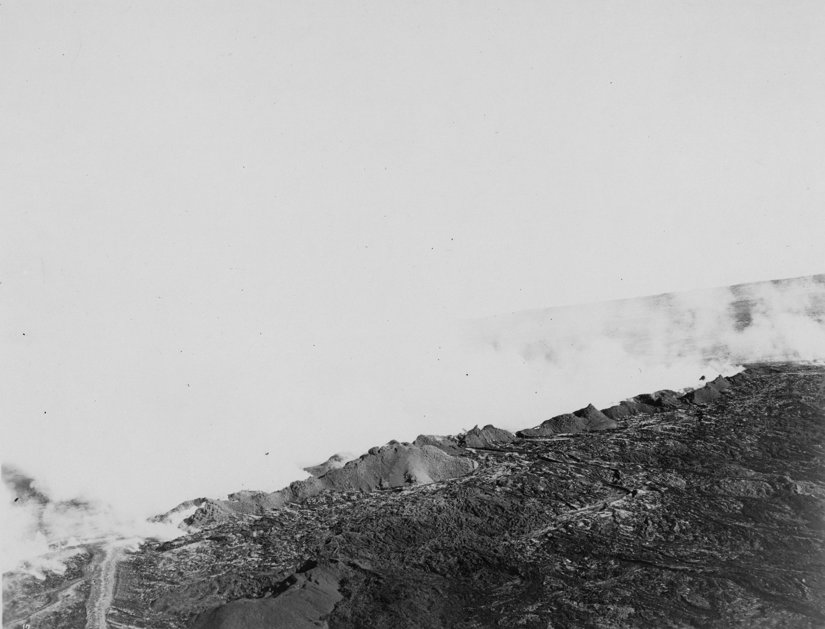 Black and white photograph of a landscape that is mostly obstructed by clouds of white smoke. Smoke takes up almost the entirety of the image and blends in with the sky, obstructing the location’s identification. The only visible piece of land is apparent at the bottom right. The land is rough and rocky and appears to be mostly dried lava.  In the bottom left corner there is a flow of lava running down out of view. A piece of elevated land can be seen in the distance towards the right side, through gaps in the cloud of smoke.