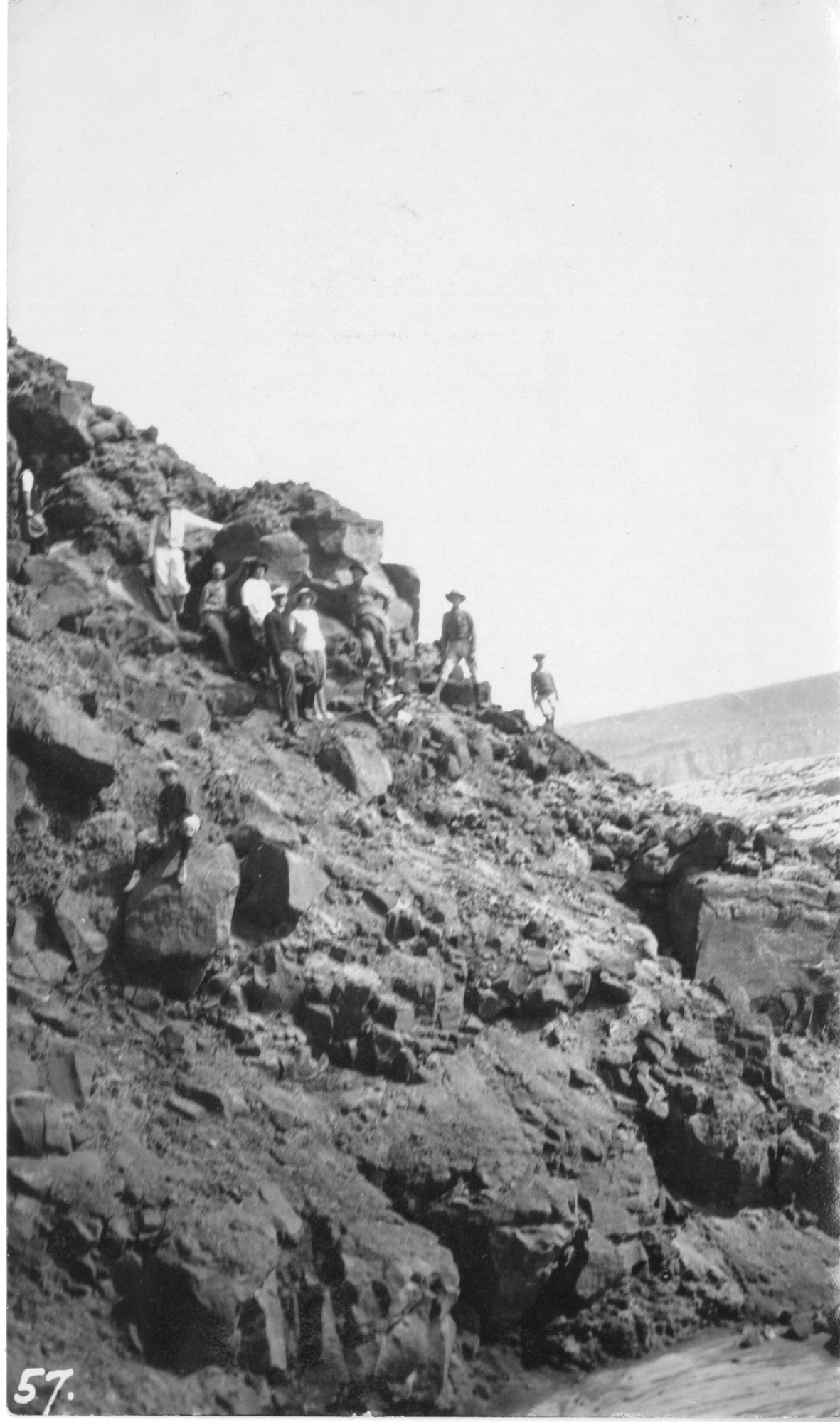 The image is in black and white. A large group of hikers in the upper left and center of the image make their way down the bluff, a steeply sloped, loose-rock incline. Huge boulders litter the area around the path, and stones jut out from the hill. The camera is far from the hikers, who are in the back of the middle ground of the image. Lava fields are faintly visible behind the incline. The sky is clear.
