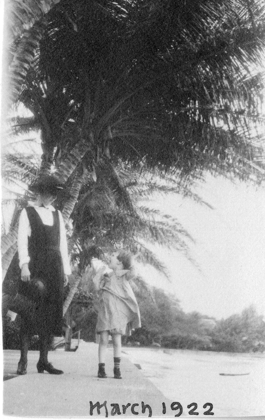 A black and white image of a woman and a child on the sidewalk near the ocean. The woman and child are located on the left side of the image. The woman is wearing a hat, a long sleeve shirt, a jumper, and heeled shoes. She is holding something in her right hand by her side. She is looking down towards the girl to the left of her. The girl is wearing a short sleeve dress and sneakers. She appears to be in motion as her dress blows from the movement. Her mouth is open wide in a big smile looking up at the woman next to her. The ocean is on the right side of the image. In the background there are palm trees visible. Along the bottom of the image, "March 1922" is written in ink.