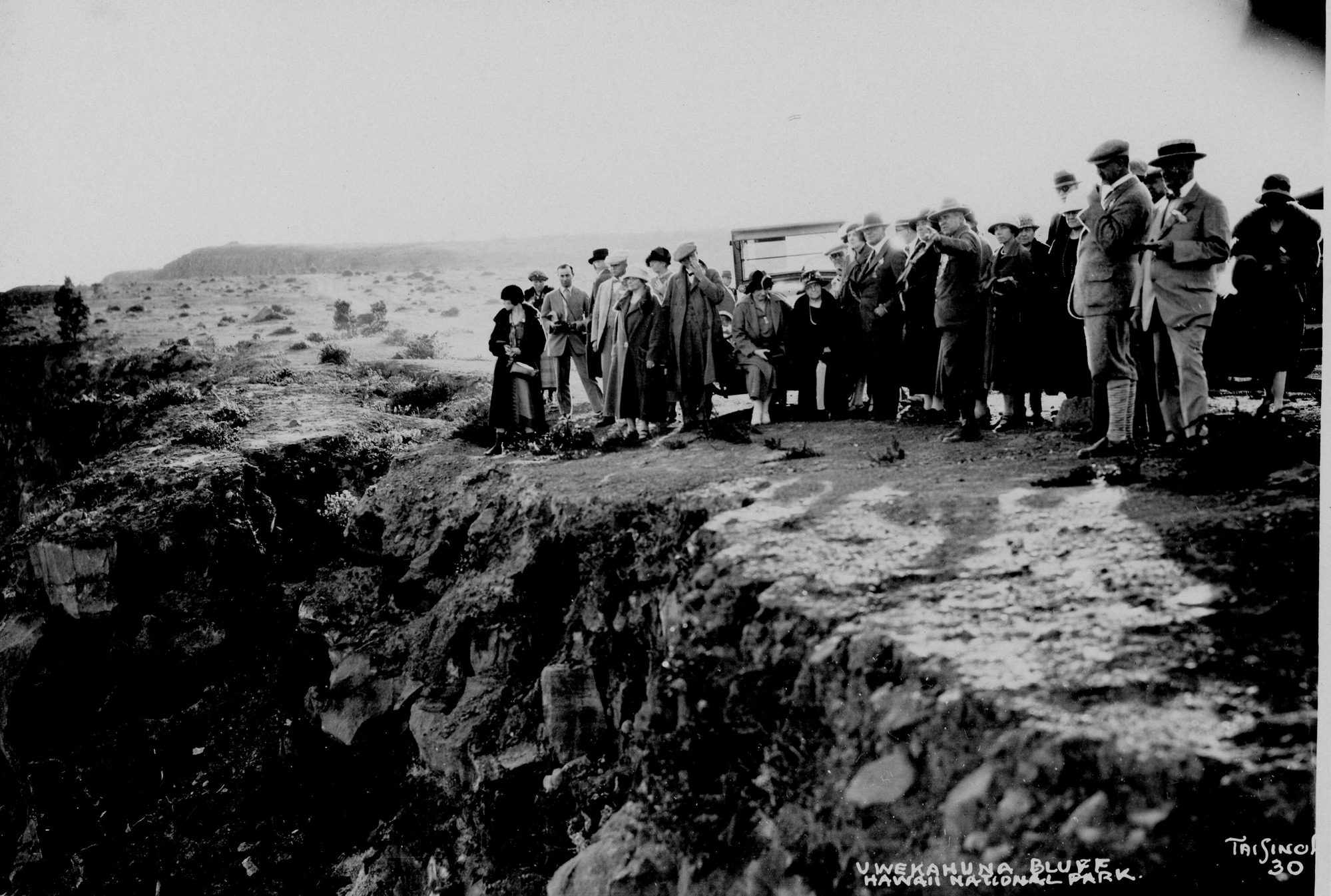 Black and white photograph of approximately twenty people standing near the edge of Uēkahuna bluff. The crowd stretches from the middle of the image to the righthand side. All are dressed in formal attire and looking out towards the left side. The rim extends from the right bottom corner of the image to the middle-left. The terrain is rocky and vegetation is sparse. Tai Sing Loo’s name is cutoff at the bottom right corner, reading “Tai Sing, 30.” Handwriting on the bottom right reads, “Uwekahuna Bluff, Hawaii National Park.”