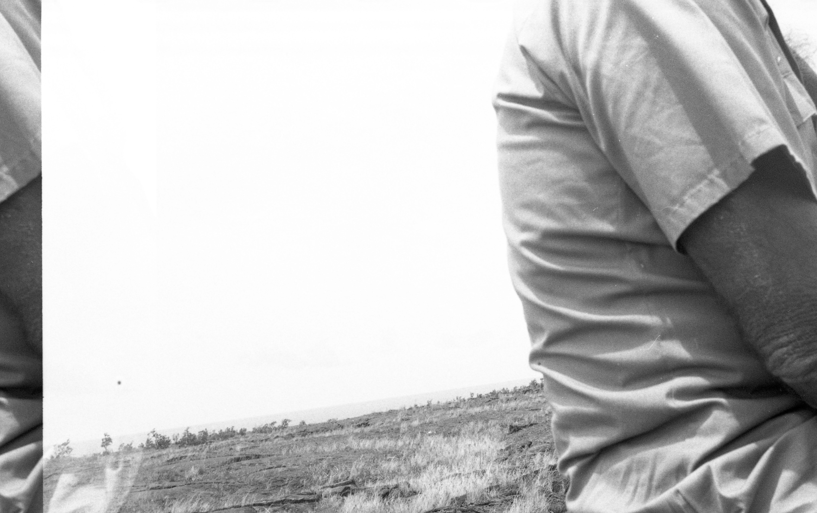 A black and white image of a flat lava field with patches of grass growing up through the cracks in the lava. On the right side of the image there is a person standing with the side of their body facing the camera. Only the person's arm, shoulder, and a portion of their back is visible.