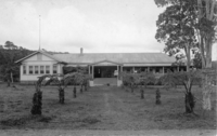 A black and white postcard of the Crater Hotel. There are palm trees lining the pathway to the front entrance of the Hotel. The entrance of the Hotel has stairs that lead up to the door. There appears to be signs in the front right of the stairs that are unreadable. There are people in dress clothes pictured on the right of the hotel sitting on the porch/lanai of the hotel. On the left side of the picture the hotel has six windows looking out to the entrance. There are also trees behind the hotel. On the right side of the picture there are ‘ōhi‘a trees in the front of the yard. There is a chimney cap on the roof towards the left side of the hotel.