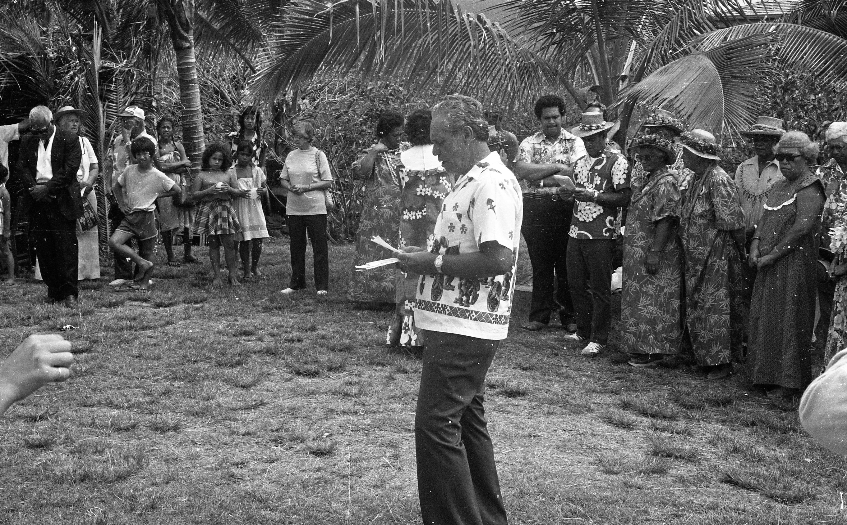 A black and white image of a man standing in a circle of cultural festival attendees reading announcements. The man is in the center of the image. His body is facing the left side of the image and is looking down at a piece of paper in his hands.  He is wearing an aloha shirt, a watch on his left wrist, and pants. There is a large group of people shoulder to shoulder behind the man reading announcements. They are all looking at him. Behind the line of people is a row of palm plants.