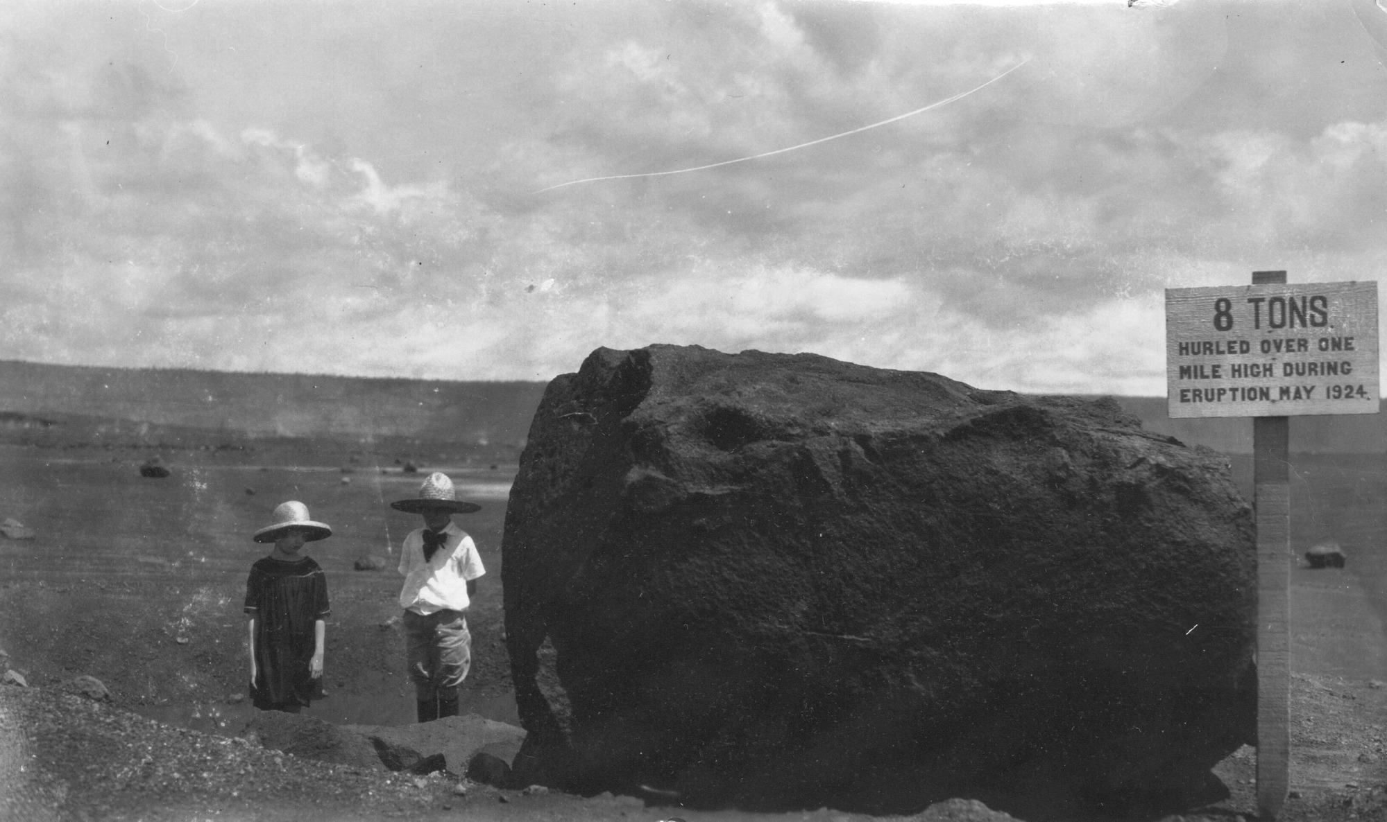 The image is in black and white. A lava bomb takes up most of the right side of the photo. It is pictured in a flat lava field. The surface of the lava bomb is rough and uneven. On the right side of the lava bomb there is a sign on a wooden post. The sign reads, “8 TONS Hurled over one mile high during eruption May 1924.” On the left side of the image standing slightly behind the lava bomb is Margaret Boles and Charlotte Lovejoy. Boles is wearing a hat and a short sleeve dress while Lovejoy is wearing a hat, a short sleeve shirt, bow tie, and shorts.