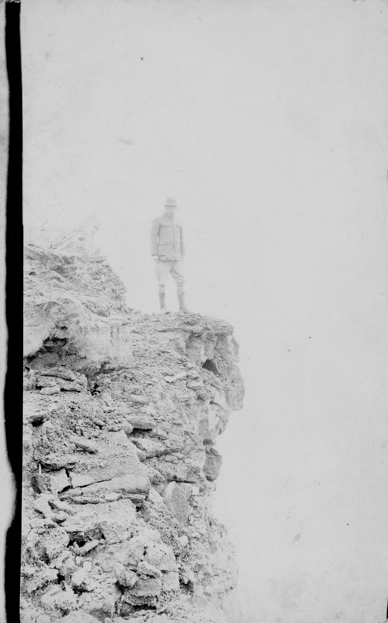 Black and white photograph of Thomas Boles dressed in National Park Service attire, standing at the edge of a rocky drop-off. Boles and the crater rim are the only things visible in the image. Boles is looking down into the crater.