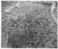 A black and white image of a flat rock surface with various petroglyph carvings covering its surface. Some of the petroglyphs are human figured. Others are triangles, circles, crosses, "X's", and lines.