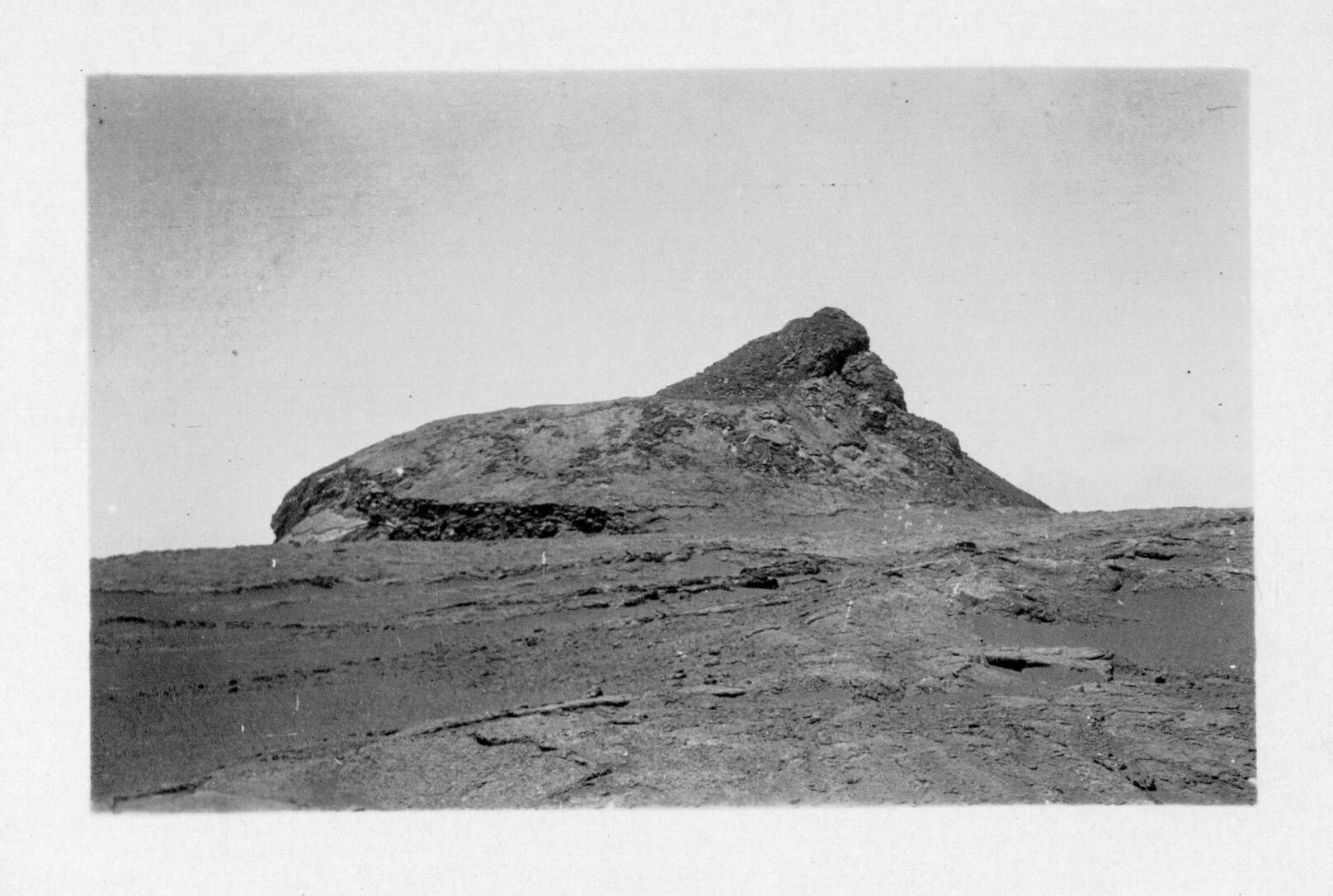 A black and white image of a large lava formation in the Kaʻu desert. The lava formation is located in a pāhoehoe lava field. It is shaped like a trapezoid in the center of the image. On the right side of the formation there is more lava on top of the trapezoid forming a high peak.