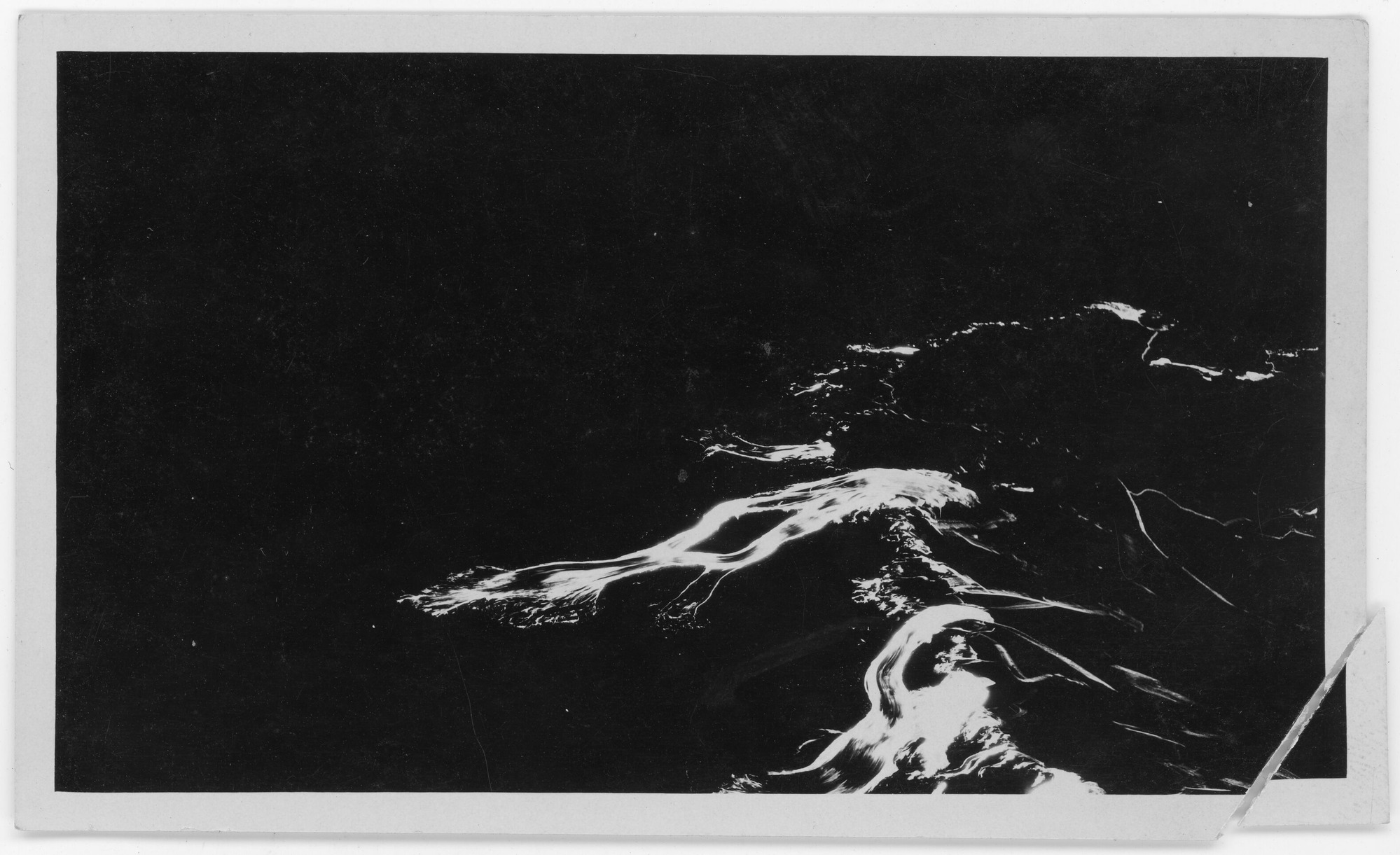 Black and white photograph of flowing lava. Lava and highlights are bright white against a black surface. The pattern of white lines take up only the lower right portion of the photograph. Some lava extends towards the middle of the photo. The lava appears to be in motion, forming a few blurred, thick pathways. There is a small tear in the photograph - the bottom right corner is unattached.
