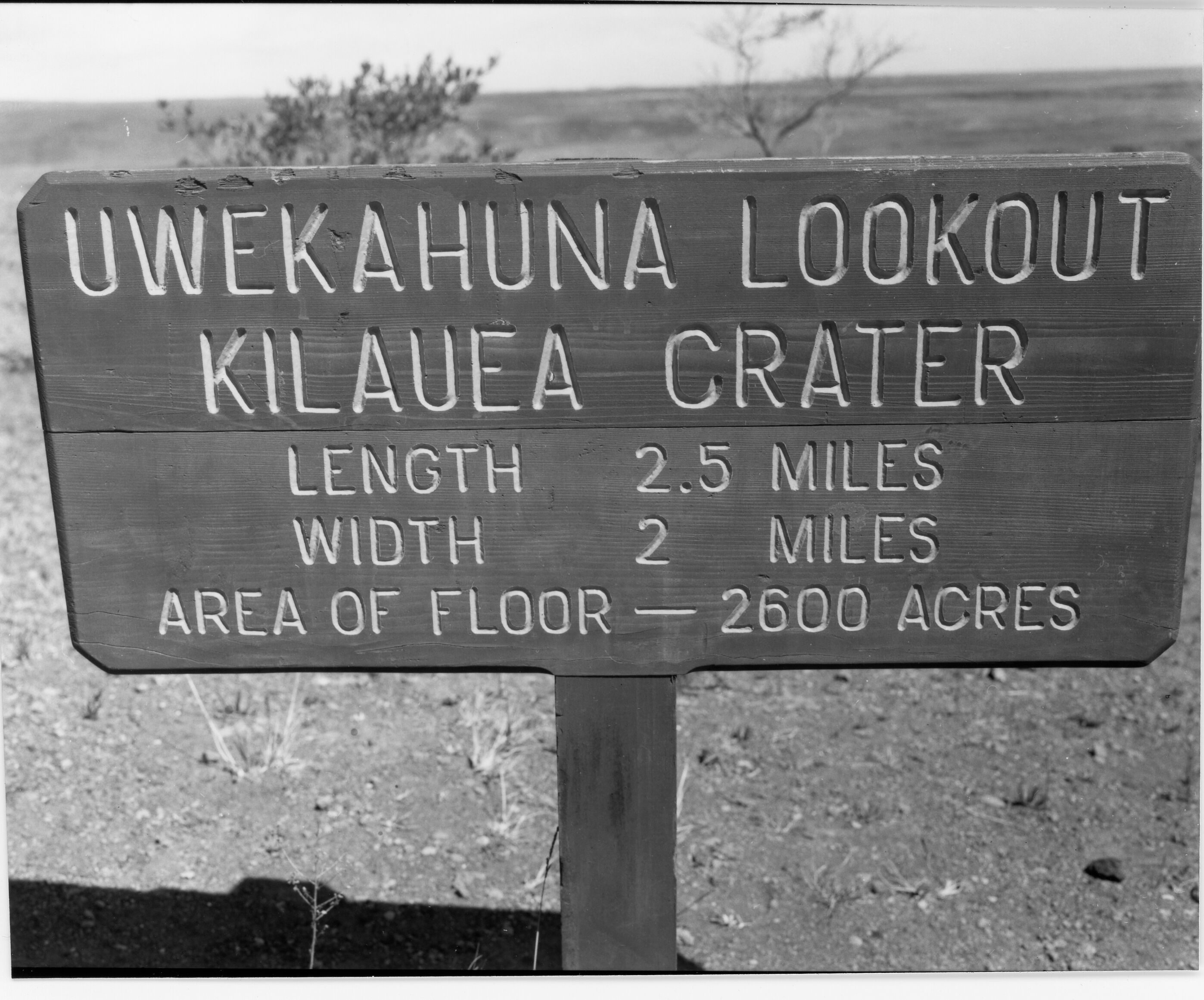 A black and white image of a wooden trailhead sign.The sign reads, "UWEKAHUNA LOOKOUT KILAUEA CRATER LENGTH 2.5 MILES WIDTH 2 MILES AREA OF FLOOR - 2600 ACRES." Behind the sign is a gravel filled landscape with a couple of trees.