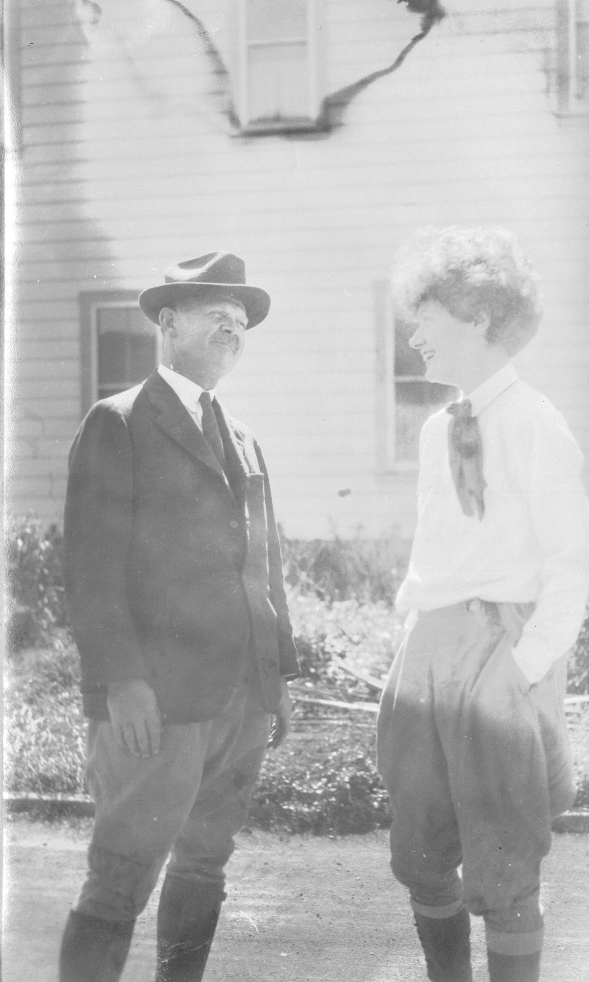 A black and white image of a man and woman standing outside of the Volcano House. The man is pictured on the left side of the image and the woman is on the right. They are turned facing each other and they are smiling. The man is identified as Thomas Boles, he is wearing a hat, a suit jacket, a white collared shirt, tie, pants, and knee-high boots. The woman is wearing a scarf, long sleeve collared shirt, pants, and knee-high socks. She has her hands in her pant pockets. In the background a curb of a road is visible followed by a side of the Volcano House.