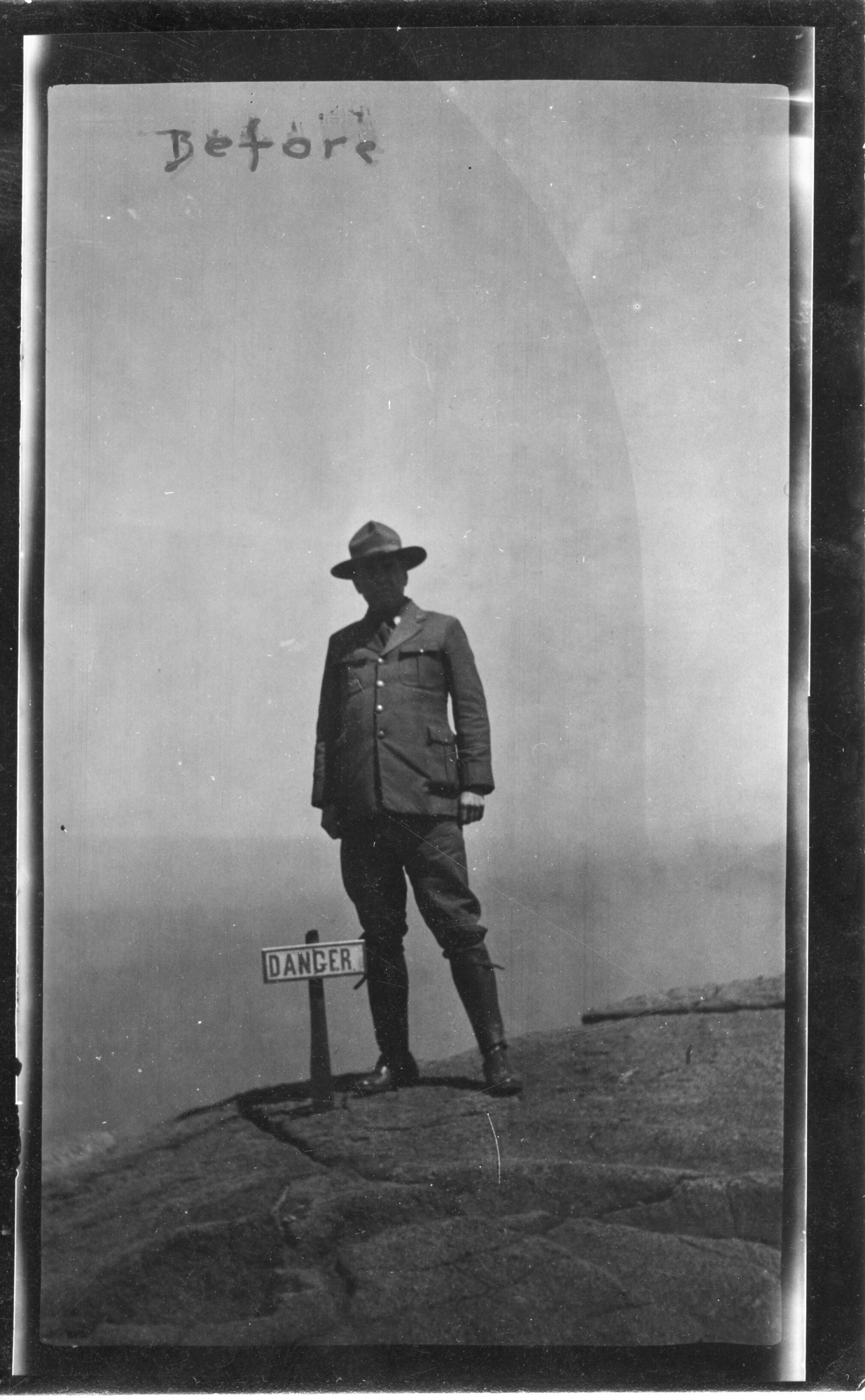 The image is in black and white. At the center of the photo is a man standing with his legs shoulder-width apart and hands at his sides. His face is obscured by shadows. He is wearing a hat, jacket, pants, and boots. To the left of his right leg is a small sign on a short post that reads, "Danger." The sign is along the edge of a crater. The ground beneath the man is composed of lava rock and is smooth and dusty. There are a few noticeable cracks running through the surface, though they are on the smaller side. The background is a blur behind the man. A black caption near the upper left of the photo reads, "Before." The writing is somewhat smudged.