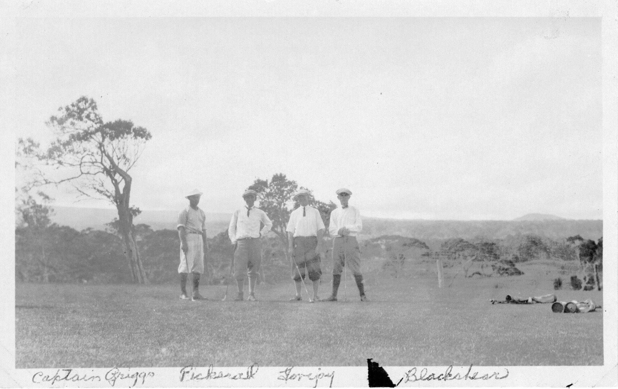 The image is in black and white. There is a dark triangular spot on the bottom white border of the image. There are four men posed on the green of a golf course, left of the center of the photo. The men are standing in a row. All of the men look at the camera, wearing flat caps, shirts, trousers, socks, and shoes. All four men are identified in the photo from the center to the left side of the image: Blackshear, Lovejoy, Pickerall, and Captain Griegs. Channing Lovejoy is the second man from the right. Behind the men, a few large trees grow, including one to the left of the group. Past these trees and a wire fence, which stretches from one end of the photo to the other, is a heavily forested area. A hill is faintly visible far off in the distance on the right edge of the photo. Label at the bottom of the image in the white border of the photo reads, "Captain Griegs, Pickarall, Lovejoy, Blackshear."