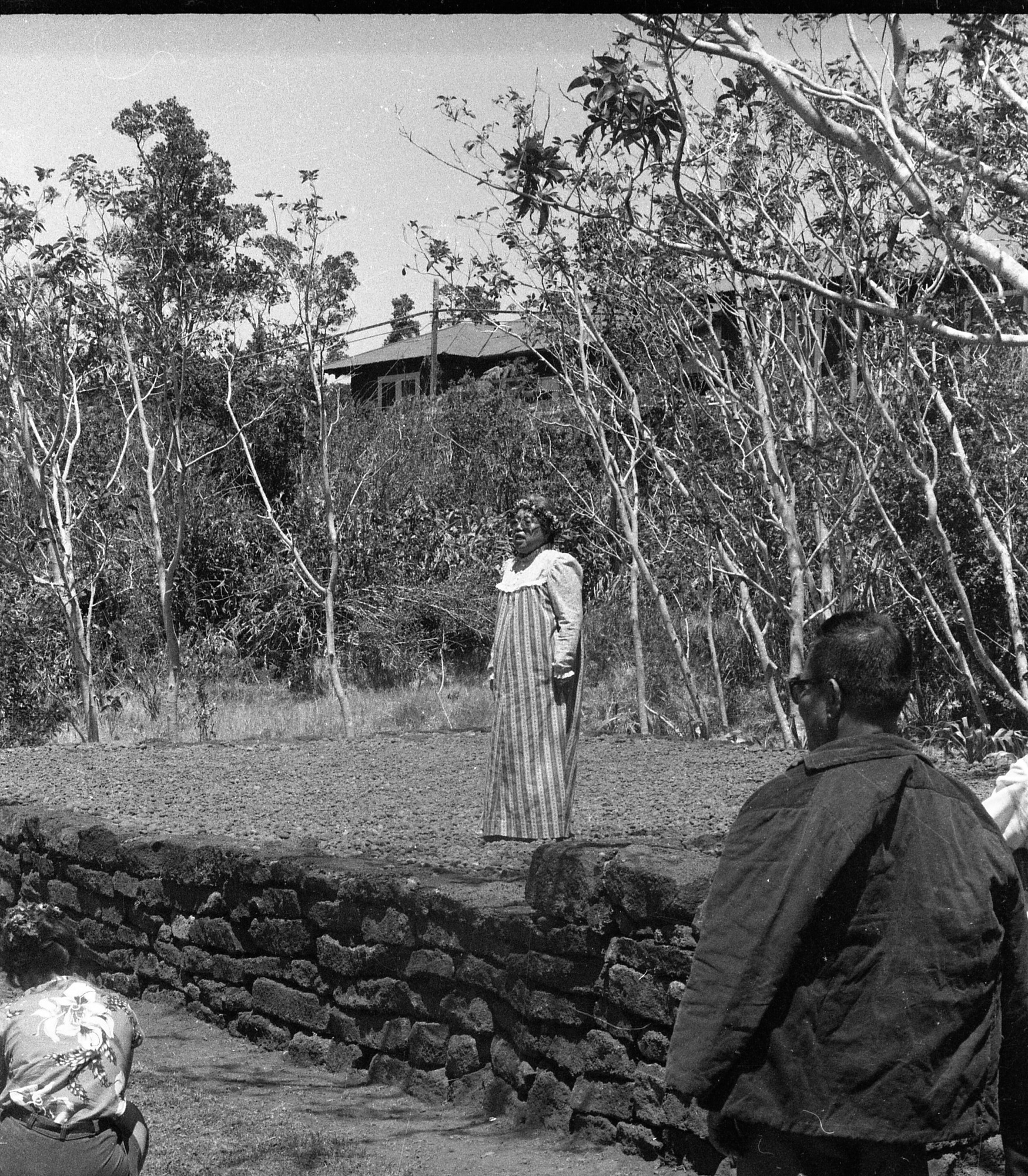 A black and white image of a woman performing on a raised platform in front of an audience.  The raised platform is bordered by a rock retaining wall. The woman is located in the center of the image. She is wearing a long sleeve dress with vertical stripes. She is looking forward towards the audience and has a floral crown on her head. In the background there is a line of trees. Beyond the trees in the upper left corner of the images is a building, the roof is visible and a part of the front. A man is located towards the right side of the image. His back is facing the camera. The man close of the image has a hoodless jacket draped around his shoulders and is wearing pants. He is watching the performer. There is a woman crouched down in the bottom left corner of the image. Her back is facing the camera. She is wearing an aloha shirt and shorts.
