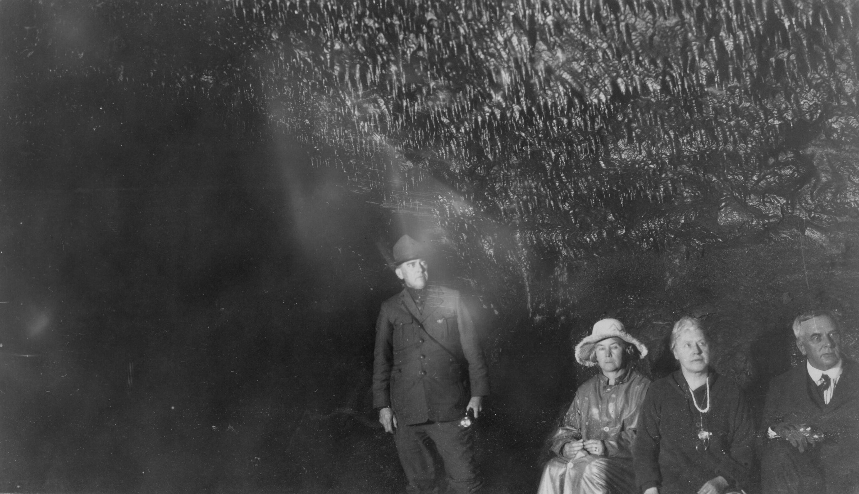 Black and white photograph of two unidentified men and two unidentified women in a lava tube. One of the men standing close to the center of the photograph appears to be dressed in National Park Service attire, holding a flashlight. The two women are seated towards the right side with the second man seated on the rightmost side of the image. The three people sitting are dressed in formal attire. All four people in the photo are looking towards the right with neutral faces. Above the group are small stalactites that cover the roof of the cave. The left side of the image is almost entirely black.