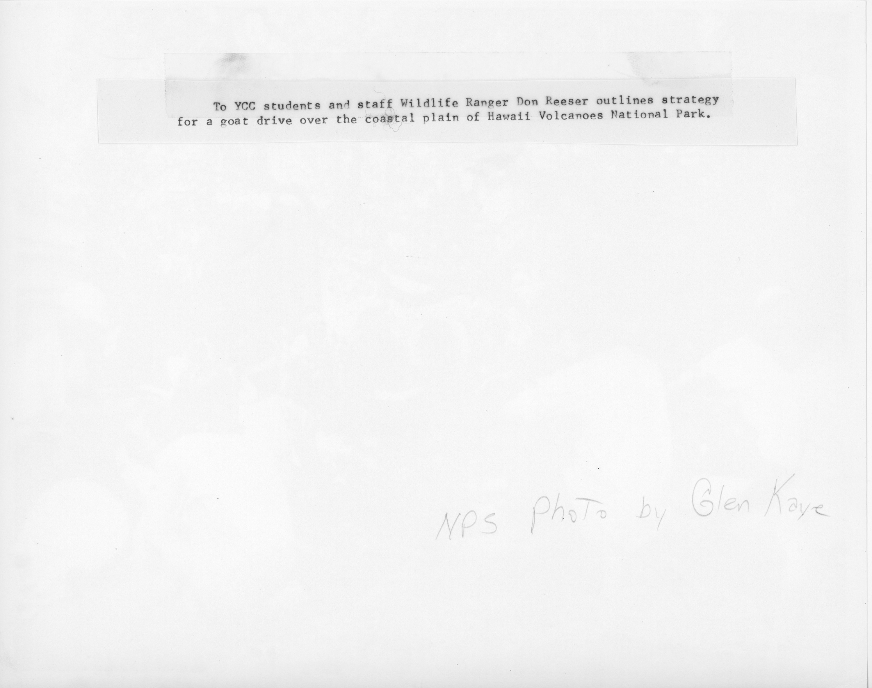 The backside of an image with Don Reeser having a meeting with Youth Conservation Corps members. At the top of the image there is a caption that reads, "The YCC students and staff Wildlife Ranger Don Reeser outlines strategy for a goat drive over the coastal plain of Hawaiʻi Volcanoes National Park.” Towards the bottom right corner of the image, “NPS Photo by Glen Kaye,” is written in pencil.
