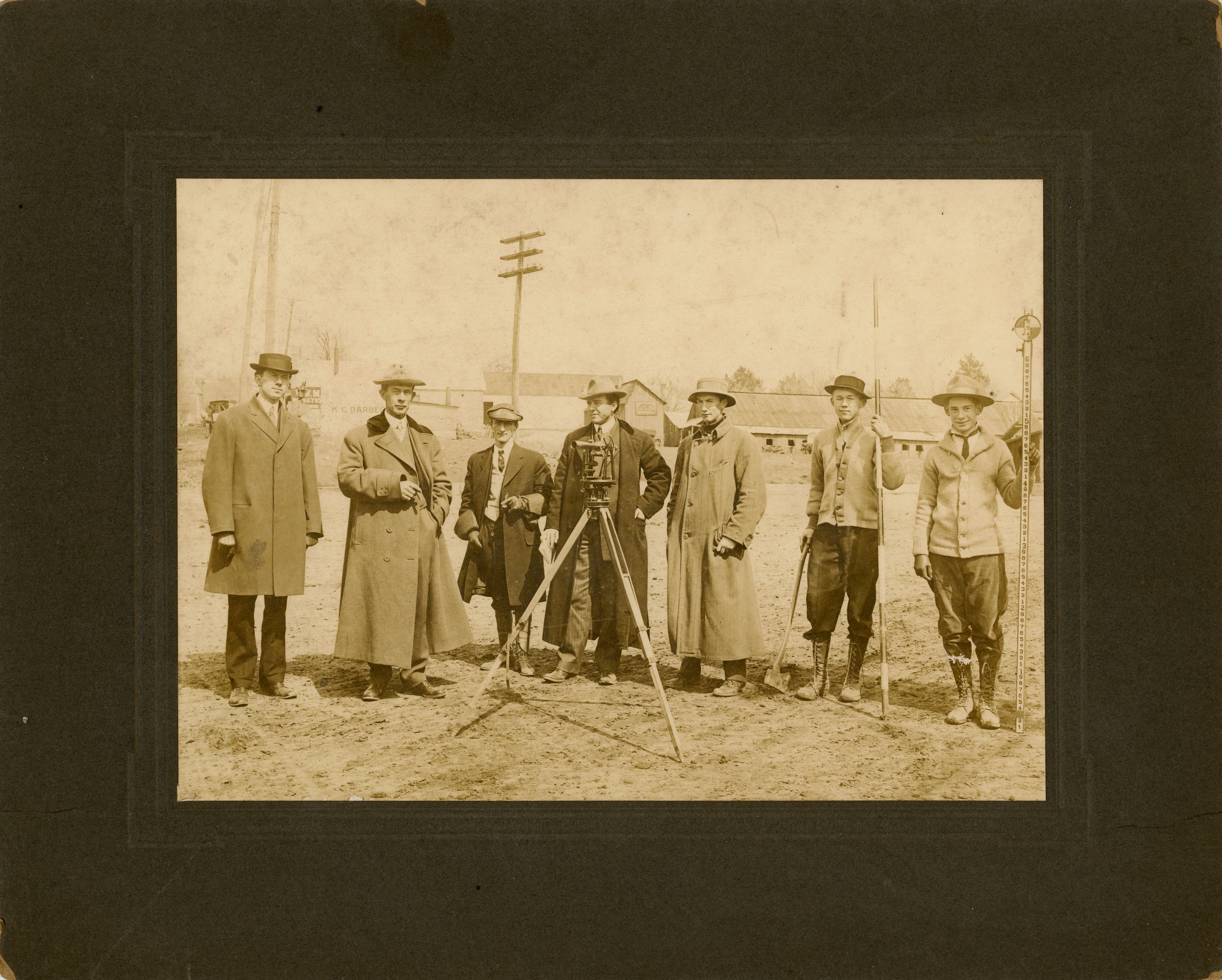 Sepia-toned, mounted photograph of seven men outdoors. A couple of men hold a cigar and a pipe, and two younger men on the right side hold long measuring equipment. A small mechanical machine stands on a tall tripod in front of the group. All of the men are dressed in formal attire with hats. Buildings and telephone poles can be seen in the background. A sign painted on one of the buildings is partially visible, reading “K. G. Barbec.” The cardboard that the photograph is mounted on is dark brown.