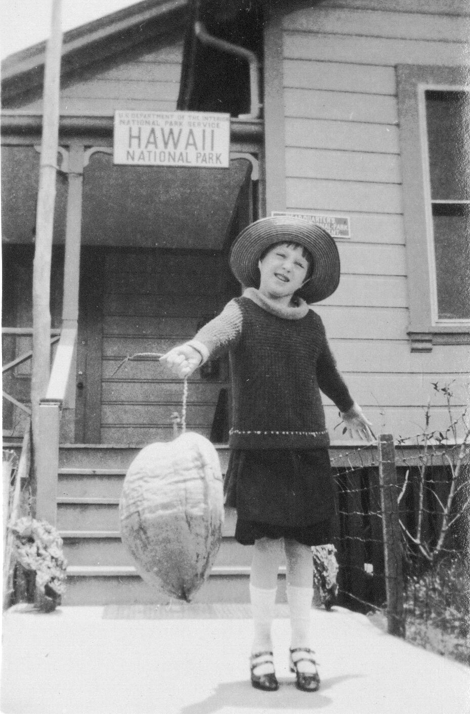 A black and white image of a girl holding a coconut outside of the old administration building. The girl is in the center of the image smiling at the camera. She is wearing a hat, a long sleeve dress, tights, and Mary Janes. Both of her arms are stretched out to her sides. She is holding a coconut by its root in her right hand out in front of her. She is standing on the sidewalk just below the staircase to the administration building. There are two signs on the building behind her. The first sign is hanging from the porch  that reads, “U.S. Department of the Interior, National Park Service, Hawaii National Park.” The second sign is partially visible behind the girl’s head, it reads, “Headquarters, Hawaii National Park, ...nt.”
