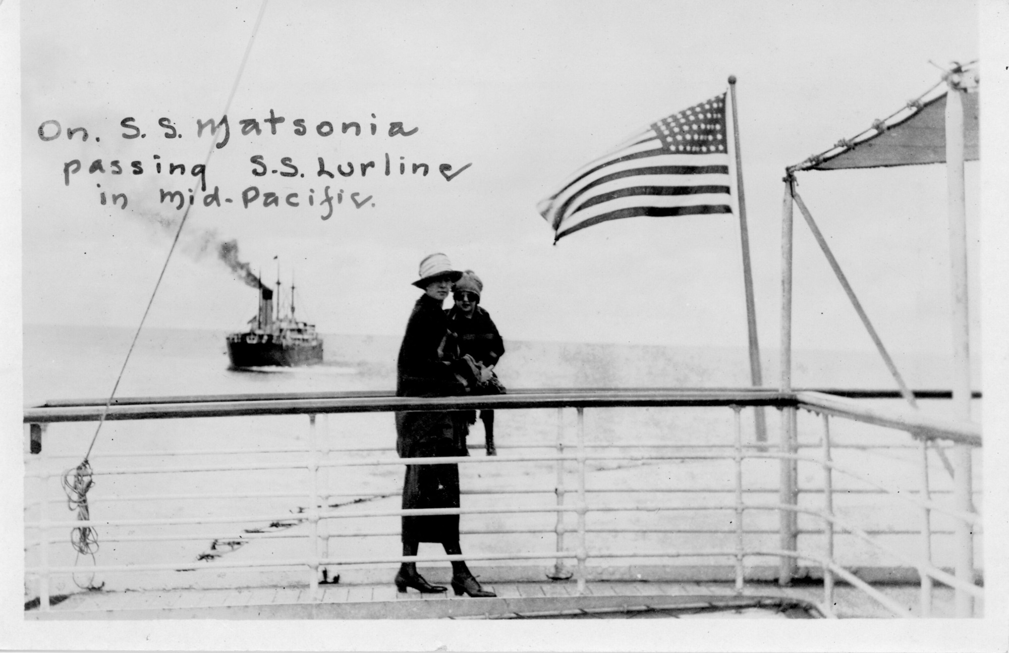 A black and white image of a woman and child standing on the deck of S.S. Matsonia. The woman and child are pictured in the center of the image standing on a walkway behind a railing. The woman is wearing a hat, coat, skirt, and dress shoes. She is looking over her shoulder at the camera. She is holding a child on top of a railing behind her. The child is wearing a beanie, sunglasses, a coat, and a dress. She is looking at the camera. On the left side of the image there is another ship in the background. Steam is rising from a tall vent on the ship. Above the ship there is a caption for the photo, “On S.S. Matsonia passing S.S. Lurline in mid-Pacific.” On the right side of the image there is an American flag attached to the railing and a covered portion of the deck.