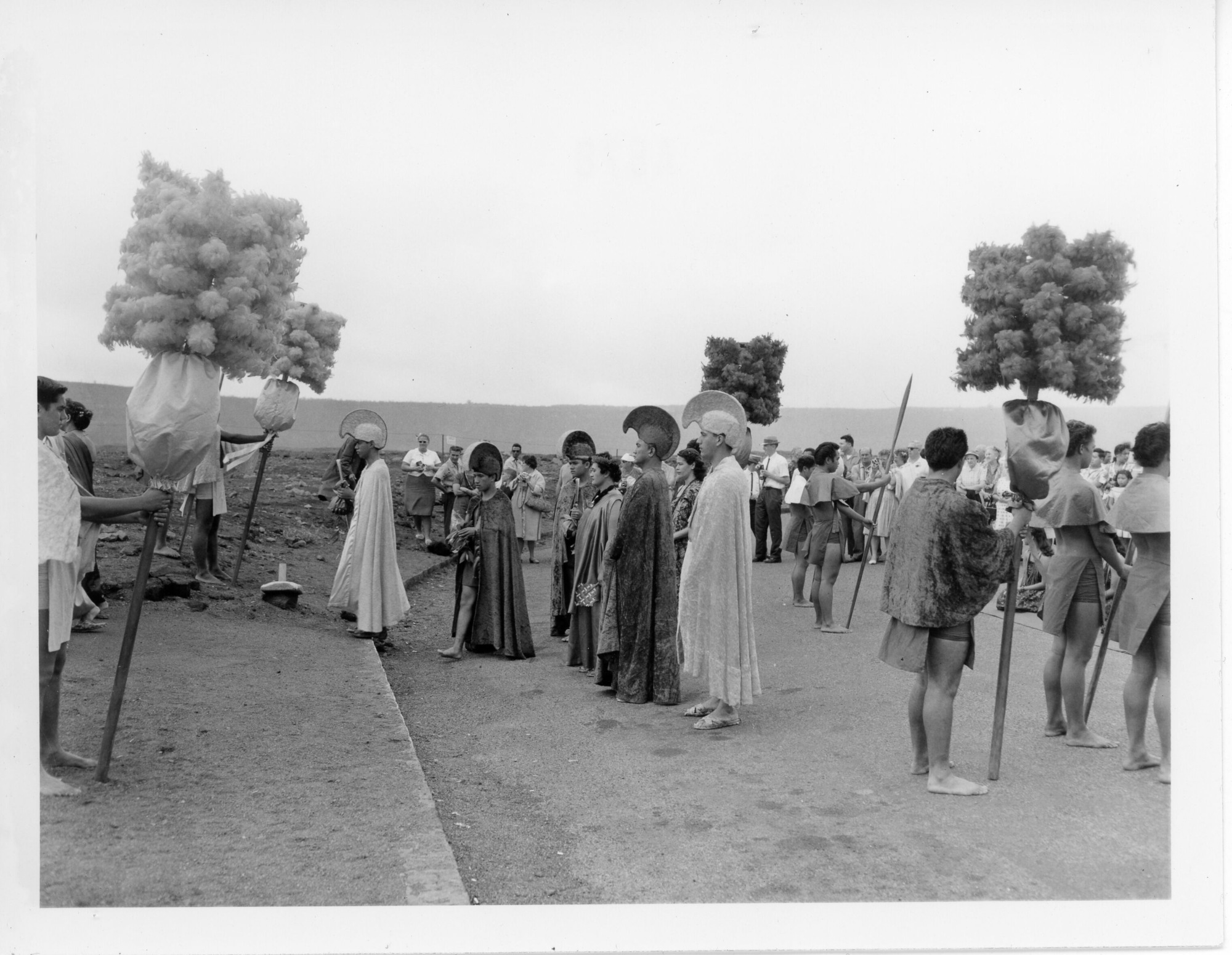 A black and white image of performers heading to the viewing area of Halemaʻumaʻu crater during a pageant. The focus of the image are five men and two women approaching the curb of a road. All seven of them are facing the left side of the image towards the curb and two men holding kāhili at the start of a trail. Three of the five men are walking in a line towards the curb. They are wearing ʻahuʻula and a mahiole and each appears to be holding something in their hands. To the left of the last man in line there is a woman. She is wearing a headband, lei niho palaoa, and a kīhei. She is holding a paper fan in her left hand. There is another woman standing behind her wearing a lei and short sleeve dress. To the left of the woman in front are two other men wearing mahiole and ʻahuʻula. Flanking this group are two more kāhili bearers wearing a ʻahu. In front of each bearer are two men holding wooden spears. All six of these men are facing the right side of the image towards a crowd watching the group.