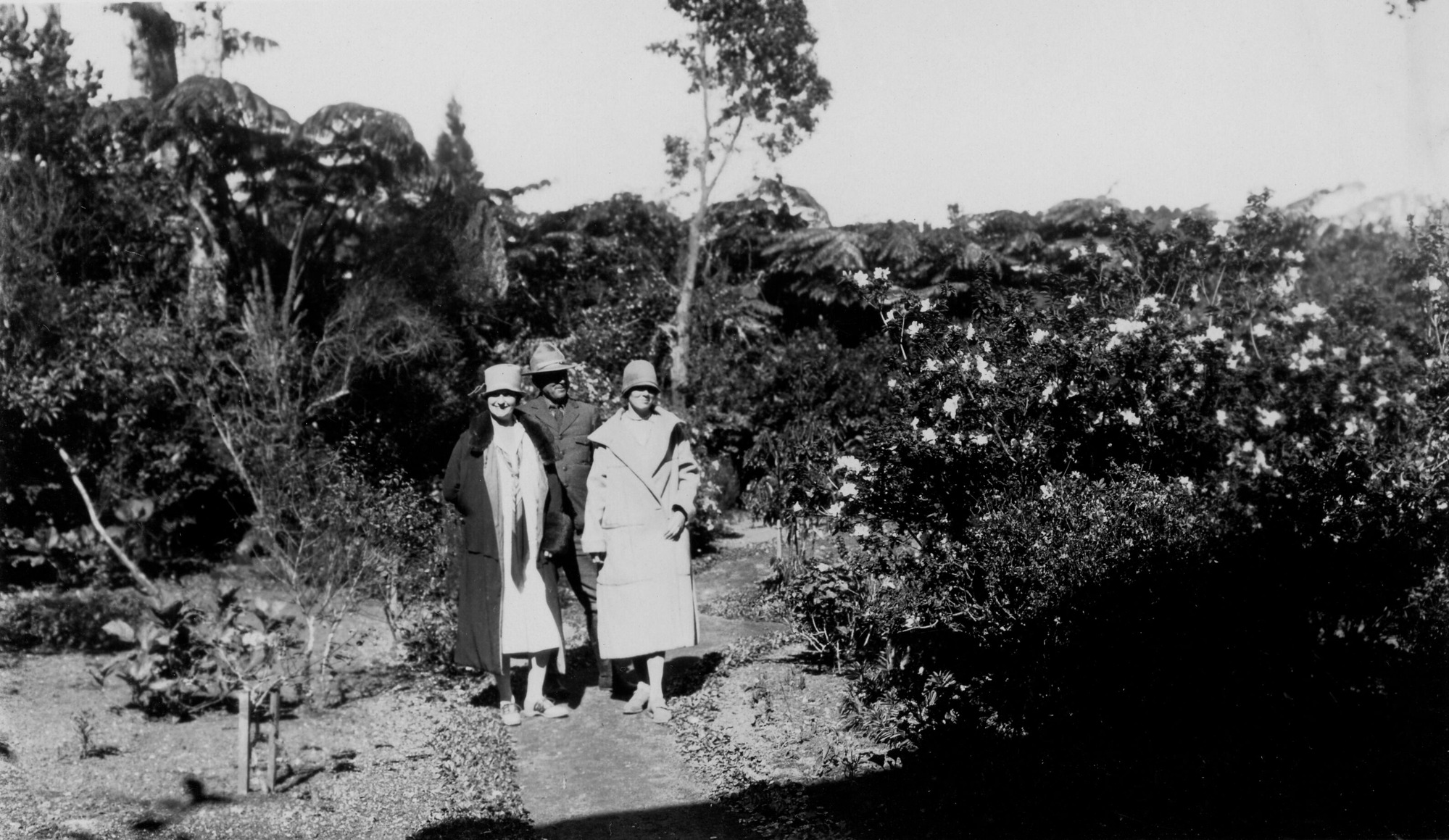 Black and white photograph of Thomas Boles, dressed in National Park Service attire, standing behind two unidentified women. All three people are smiling and facing the camera. They stand in the middle of a path to the left of the center among lush vegetation. A large bush with brightly colored flowers is present to the right side.