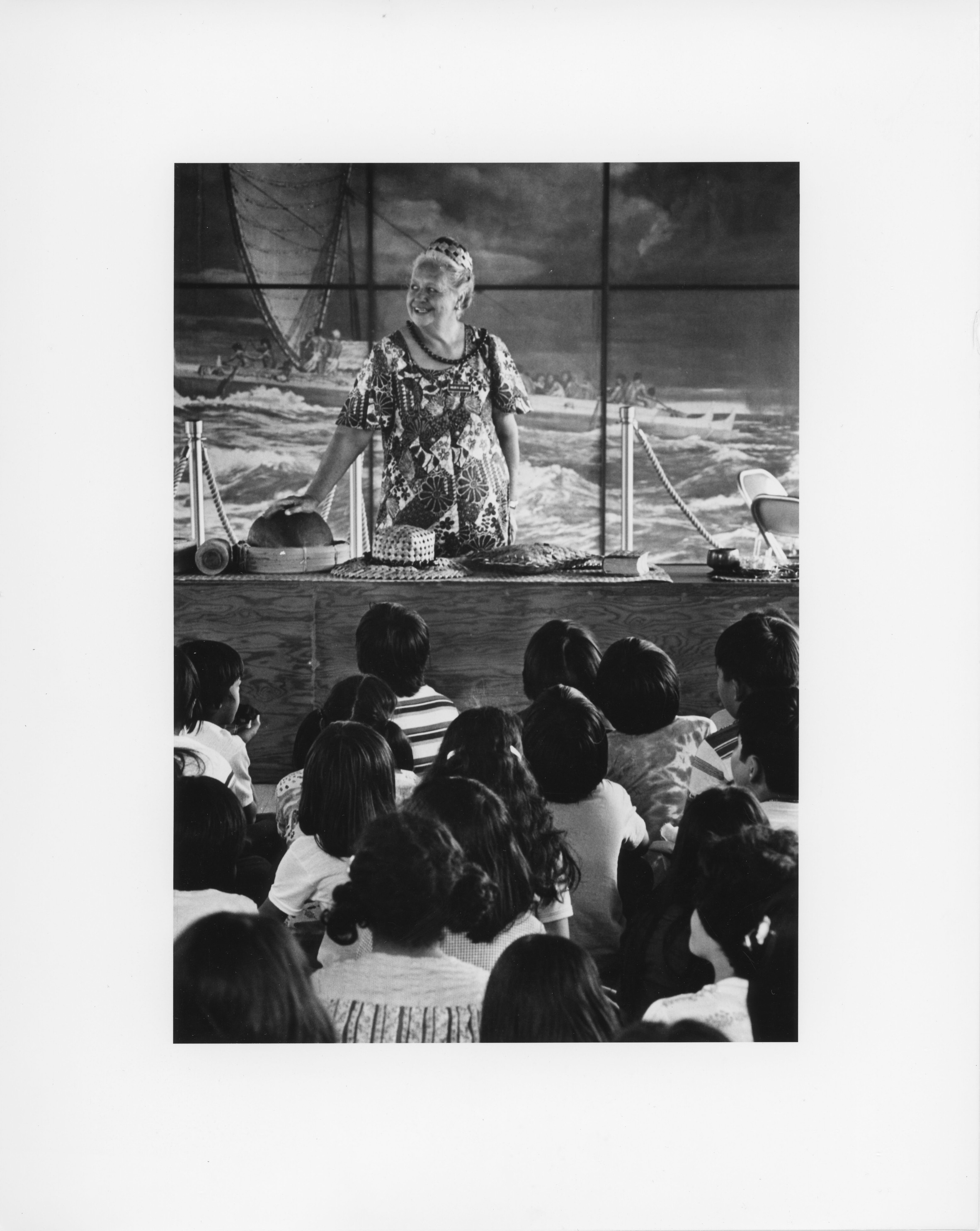A black and white image of a woman named Helen Lee Hong, presenting to a group of children at the Wahaʻula Visitor Center. Helen is standing behind a table. She is wearing a headband, a lei, and a short sleeve shirt with a tropical leaf print. She is looking towards the left side of the image. Her right hand is placed on top of a round object on the table. There are various objects spread out on the table including a woven hat, bag, book, and a calabash or bowl. Behind her is a large graphic image of a traditional Hawaiian outrigger canoe with passengers on it. There is a rope barrier in front of the image. In front of the table taking up the bottom half of the image is a large group of children sitting down watching Helen. Their backs are towards the camera.