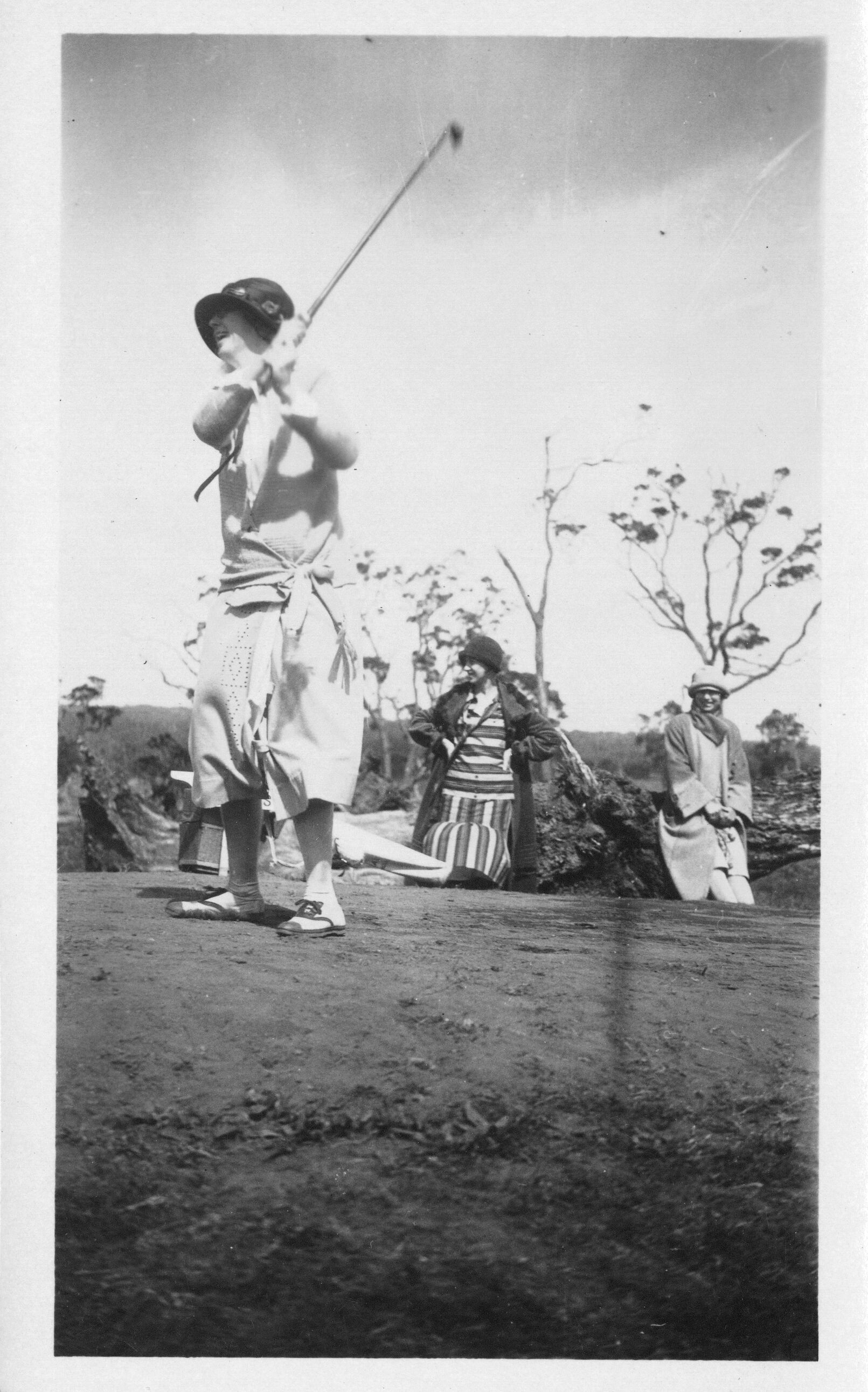 A black and white image of a woman swinging a golf club. The woman is pictured on the left side of the image standing on a raised tee mound. She is facing the left side of the image wearing a hat, a short-sleeve shirt, a sweater tied around her waist, a skirt, and sneakers. Her right arm is across her chest holding onto the end of a golf club with both of her hands. The golf club is swung over her left shoulder. She is smiling looking into the distance towards the left side of the image. Behind her there is a golf bag. There are two other women below the tee mound watching the woman swing the golf club. They are both wearing hats, long coats, and dresses.
