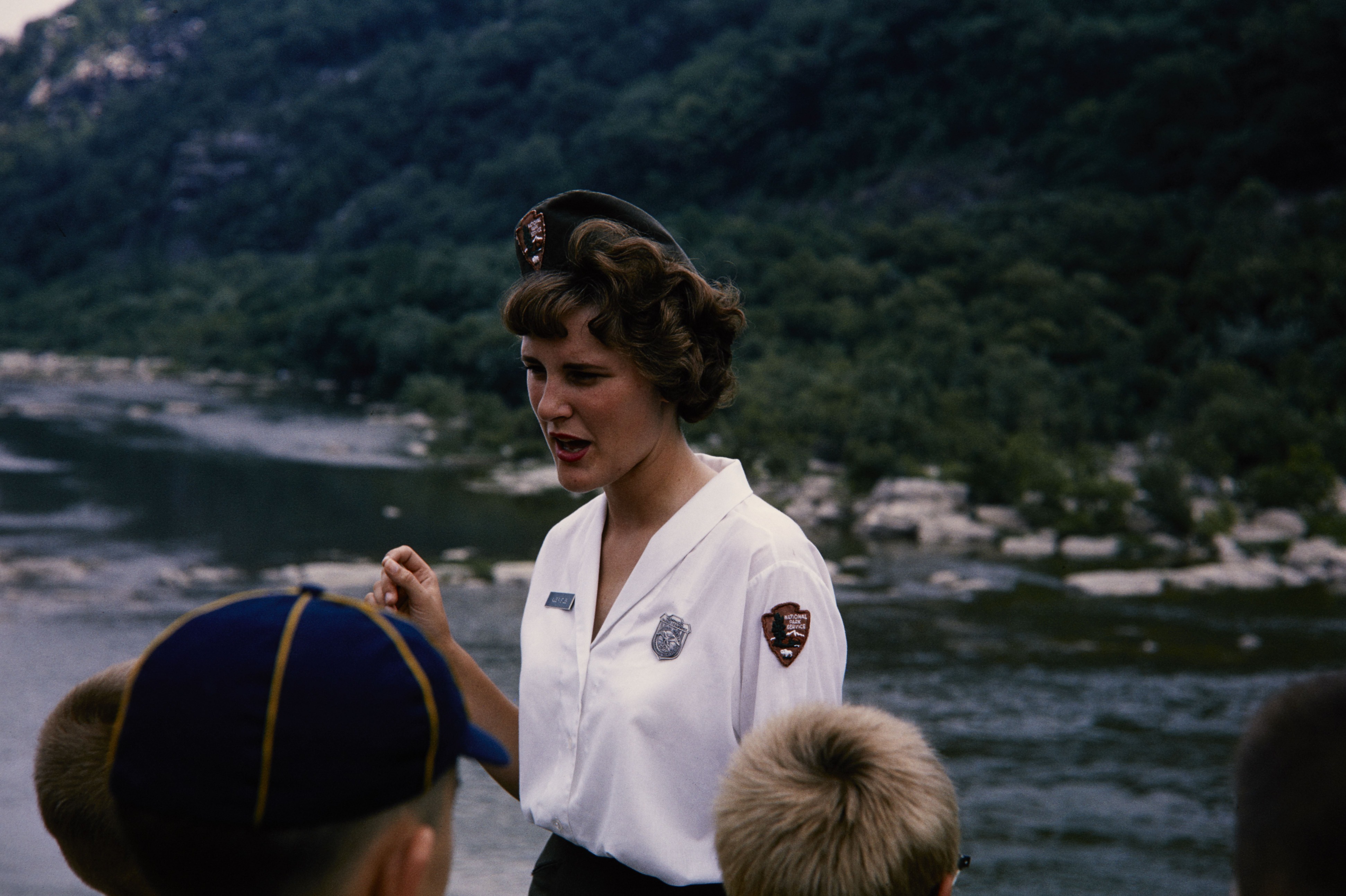 Woman interpreter in NPS uniform speaks to a group of boy scouts along a river.
