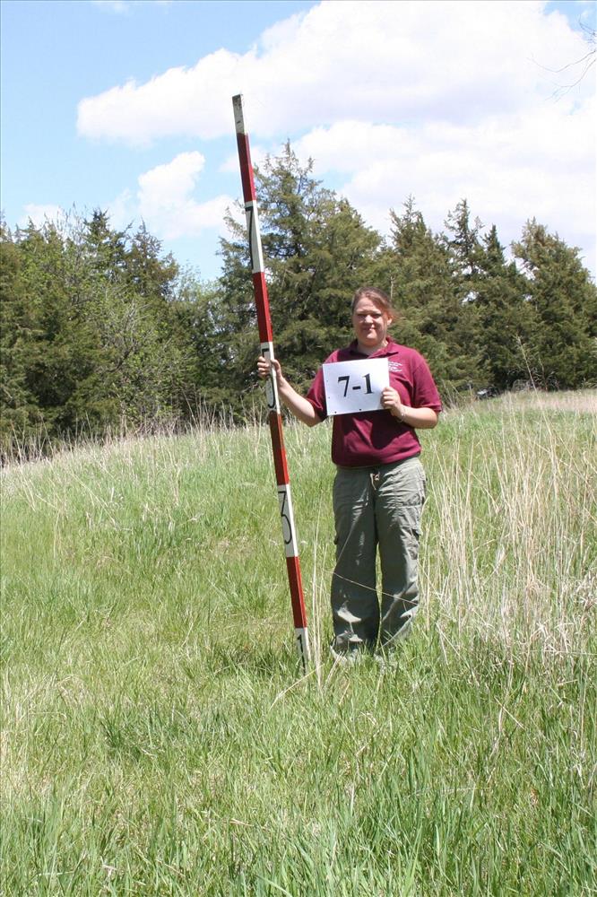 A SCA intern is holding a stadia rod and a sign in the prairie.