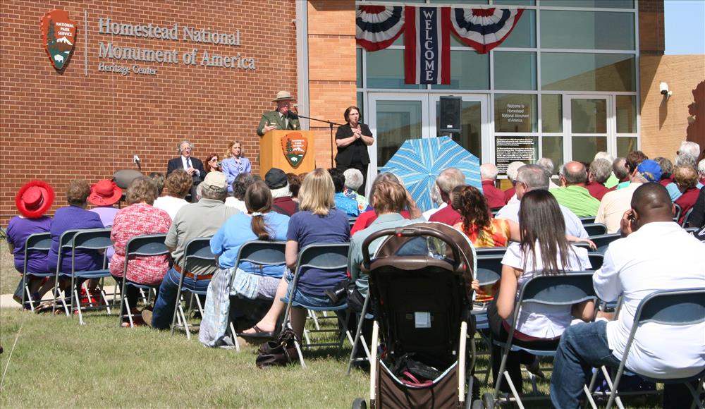 A crowd of people sitting outside in front of a stage. Mark Engler, Superintendent, is speaking at the podium with a sign language interpreter to the right.