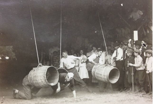 3 empty barrels are hung from the ceiling with young men attempting to dive through them. 