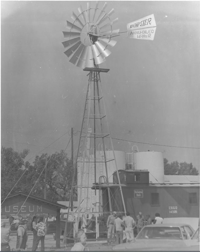 A 14 foot No. 12 Dempster windmill. There are many people near the base of the tower. There appears to be an event. A Burlington train car and a structure labeled Museum are next to the windmill.