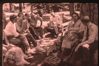 A smiling woman in American Indian attire sits with woven baskets in a demonstration. A male park ranger sits to her left.