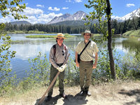 A ranger is holding an old broken post marker and a volunteer stands beside him. They are in front of Manzanita Lake and Chaos Crags is prominent in the background and part of Lassen Peak is visible to the right.