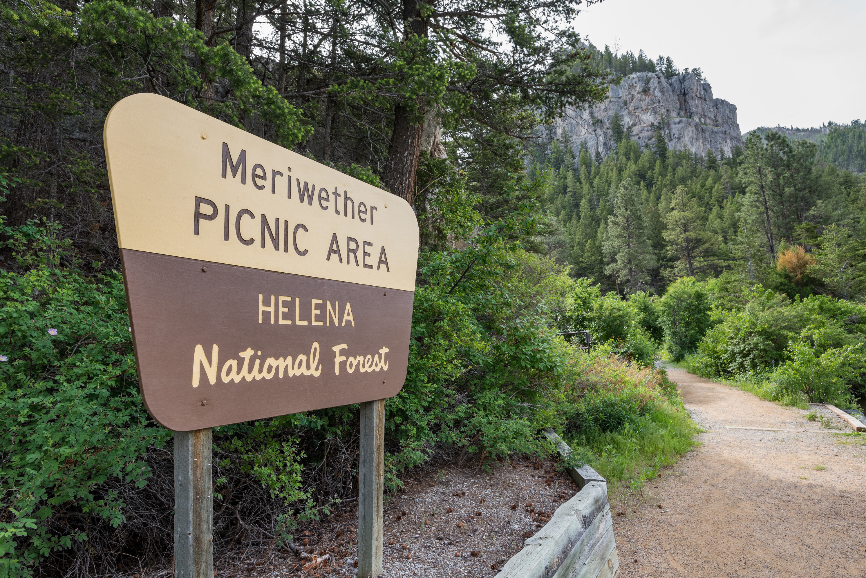 Sign reads: Meriwether Picnic Area Helena National Forest. Trail winds past sign with grey cliffs and forest in the distance