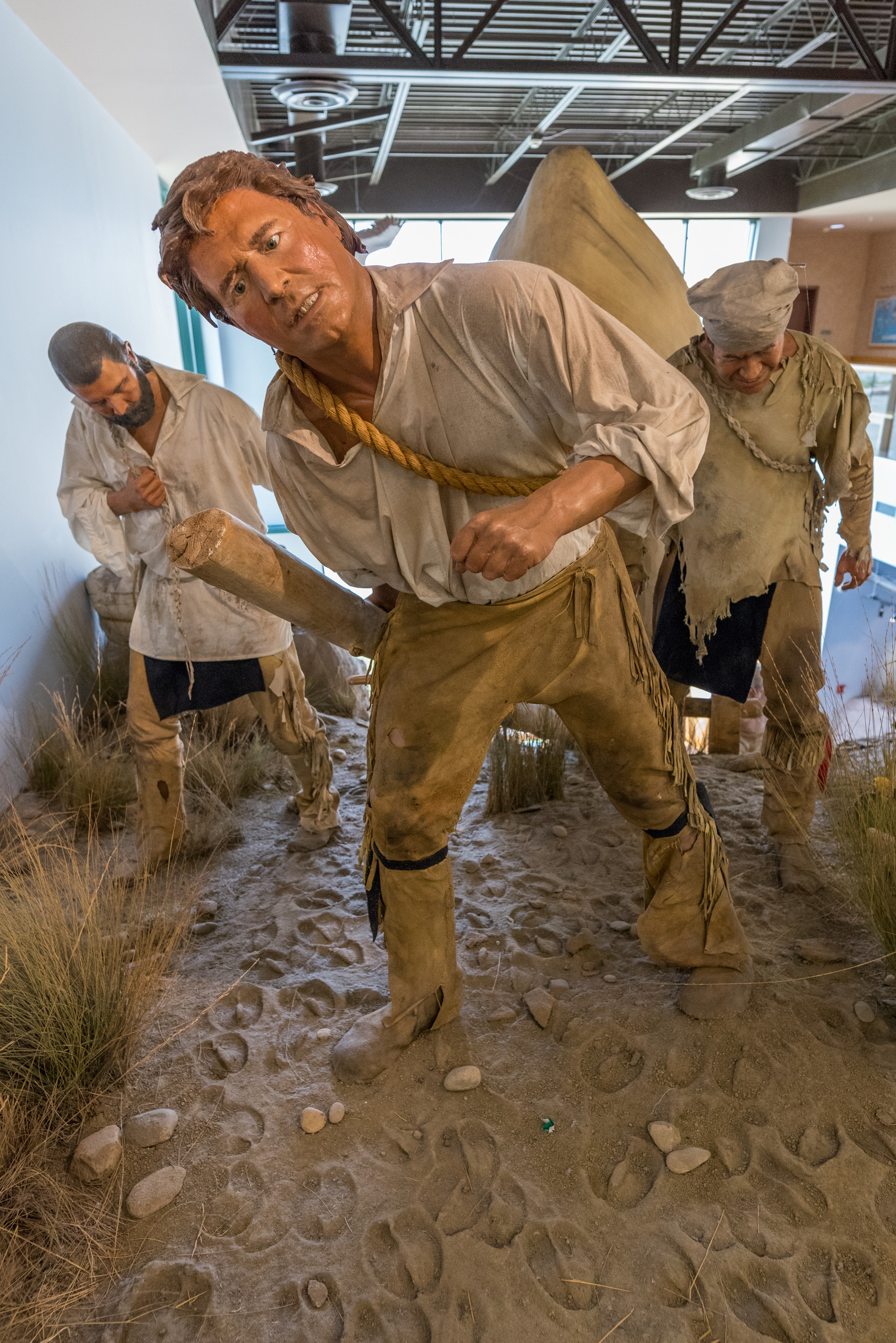 Sculpture of three men in period clothing grimacing while portaging boat through mud