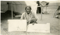 Big Beaver Seated in Front of a Tent Holding Two Pieces of His Artwork