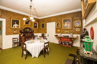 Room with dining table set for four at center with white tablecloth and wooden chairs. Portrait paintings with gold frames hang on the walls with gold floral wallpaper over white wood paneling. A red altar table with gold decorations holds vases and a plate.