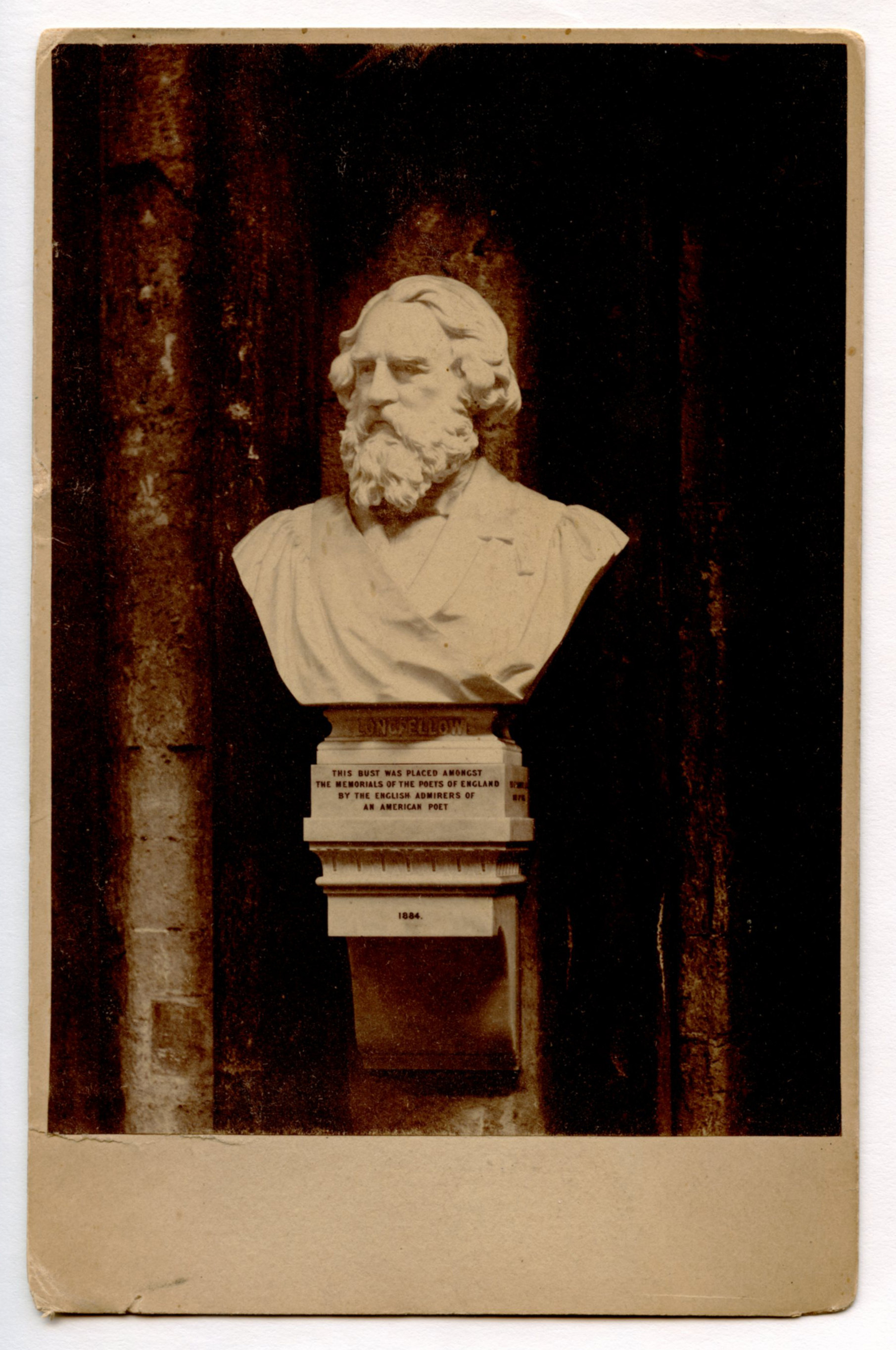 Photograph of Henry Wadsworth Longfellow portrait bust carved in white material. Bust is attached to a stone wall. There is a pedestal beneath it also attached to the wall. Longfellow looks to be wearing a graduation gown. His hair and full beard are carved in detail.