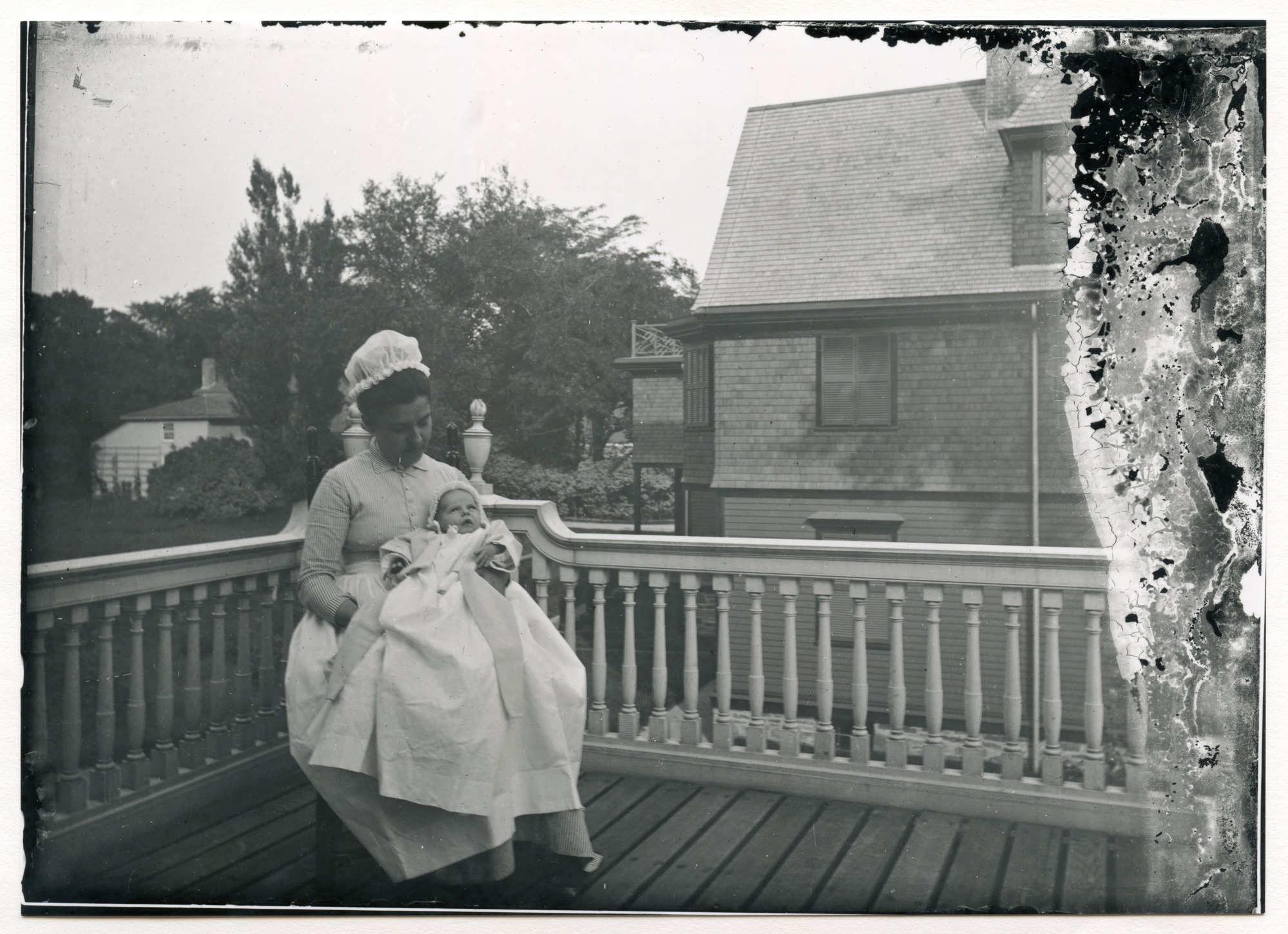 White woman in tall white head covering sits and holds a young white baby on her lap on an upper floor balcony. The neighboring house is visible to the right. Some kind of carriage house is visible to the left. Trees in background.