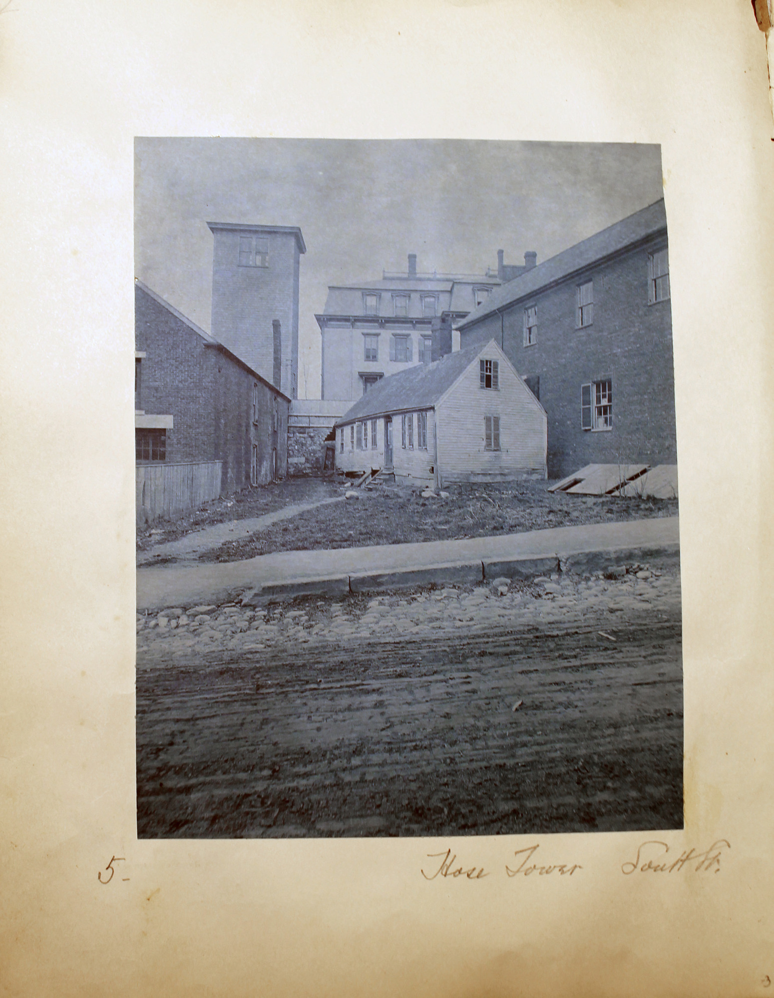 Blue toned photo of unpaved street with small dwelling in center and taller square structure to left.