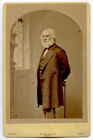 Image shaped in a rounded arch. Almost a full-length portrait of Henry Wadsworth Longfellow standing at three-quarter profile to his right. He holds a cane behind him. A window and column or grandfather clock is in the hazy background to the left. Longfellow has chin-length white hair and a full-length white beard.