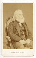 Henry Wadsworth Longfellow sits in an upholstered arm chair with his hands in his lap against a plain background. He faces his left. His vest and coat are dark, and his trousers are thinly striped. Longfellow has chin-length white hair and a full white beard.