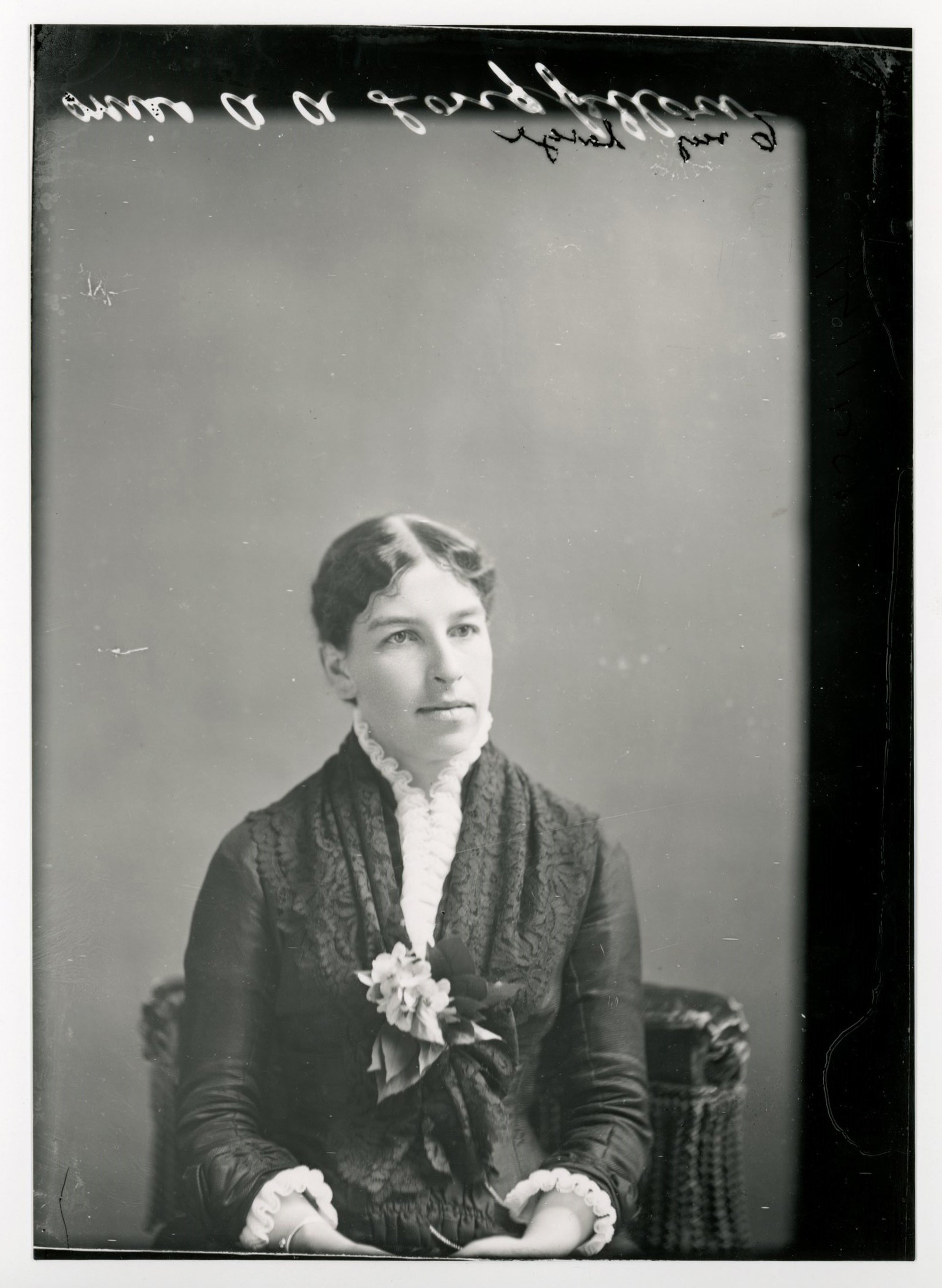 A young dark-haired white woman sits in a tasseled chair with her hands in her lap. She wears a dark-colored dress with white cuffs and a white neckline.